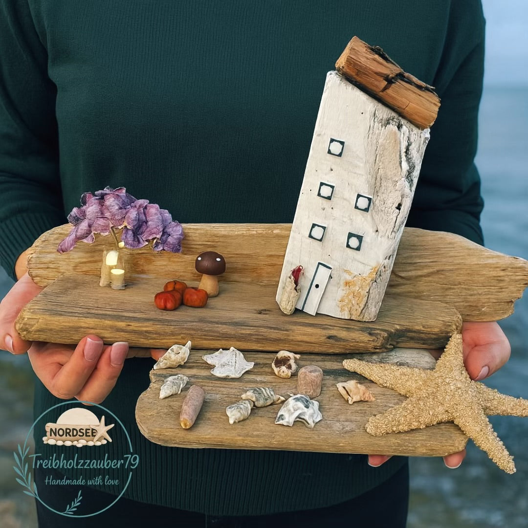 Maritime Deko aus echtem Treibholz mit einer Holzhaus-Skulptur und herbstlicher Deko als Küstenmotiv in den Händen gehalten.