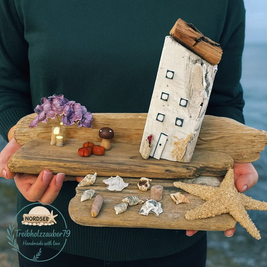 Maritime Deko aus echtem Treibholz mit einer Holzhaus-Skulptur und herbstlicher Deko als Küstenmotiv in den Händen gehalten.