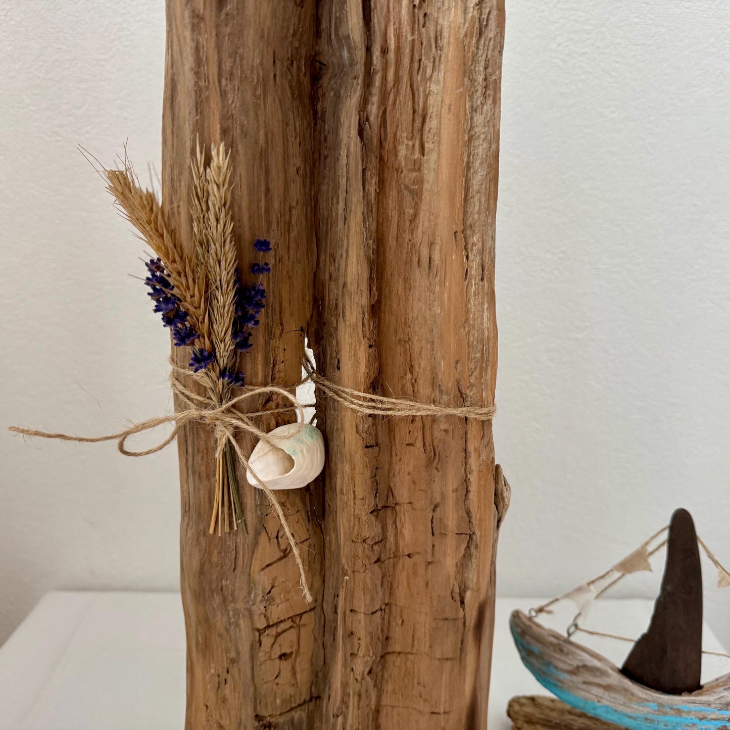 Rustikale Treibholzskulptur, verziert mit einem Bündel getrocknetem Weizen und lila Blumen, mit Bindfaden gebunden.
