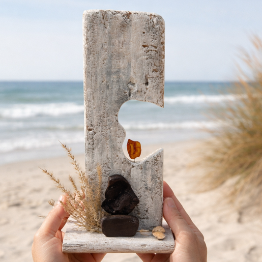 Skandinavische Bernstein Deko aus Treibholz wird am Strand mit Meer und Dünen im Hintergrund in den Händen gehalten