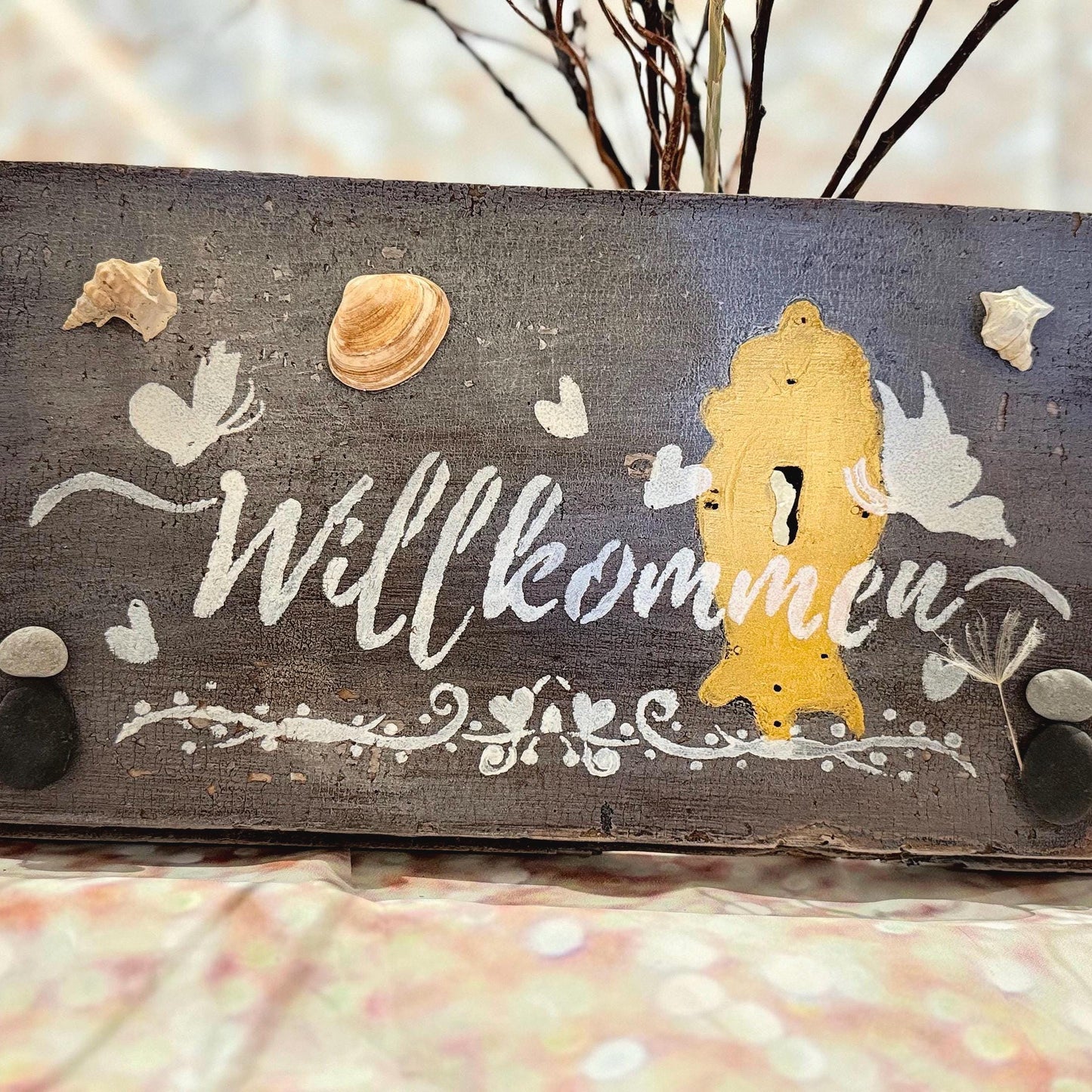 Ein rustikales Holzschild mit dem Wort "Willkommen" in weißer Schrift. Das Schild ist mit Muscheln, Schmetterlingen