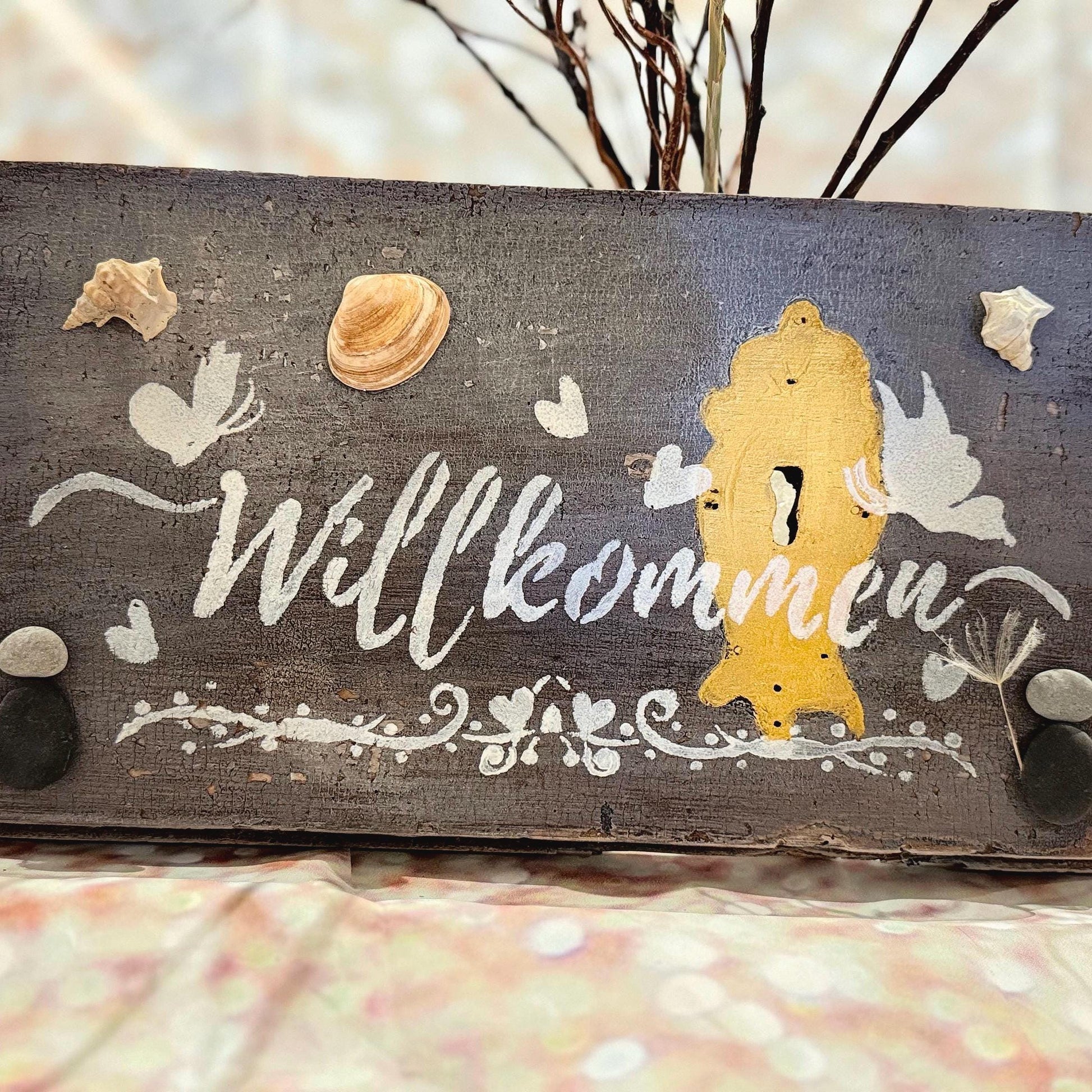Ein rustikales Holzschild mit dem Wort "Willkommen" in weißer Schrift. Das Schild ist mit Muscheln, Schmetterlingen