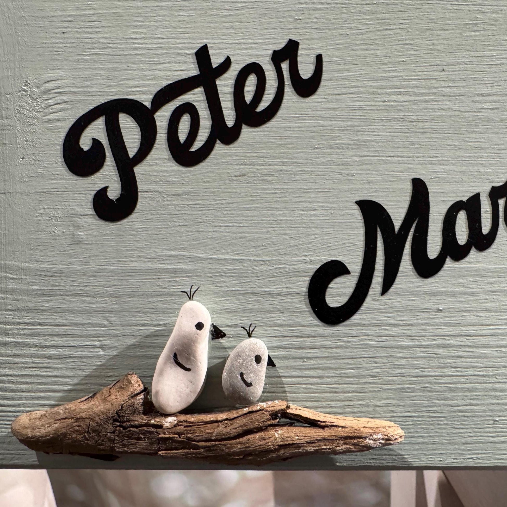 Hellblaues Holzschild mit den Namen "Peter" und "Marie" in schwarzer Schrift. Zwei kleine Steinvögel mit lächelnden Gesichter