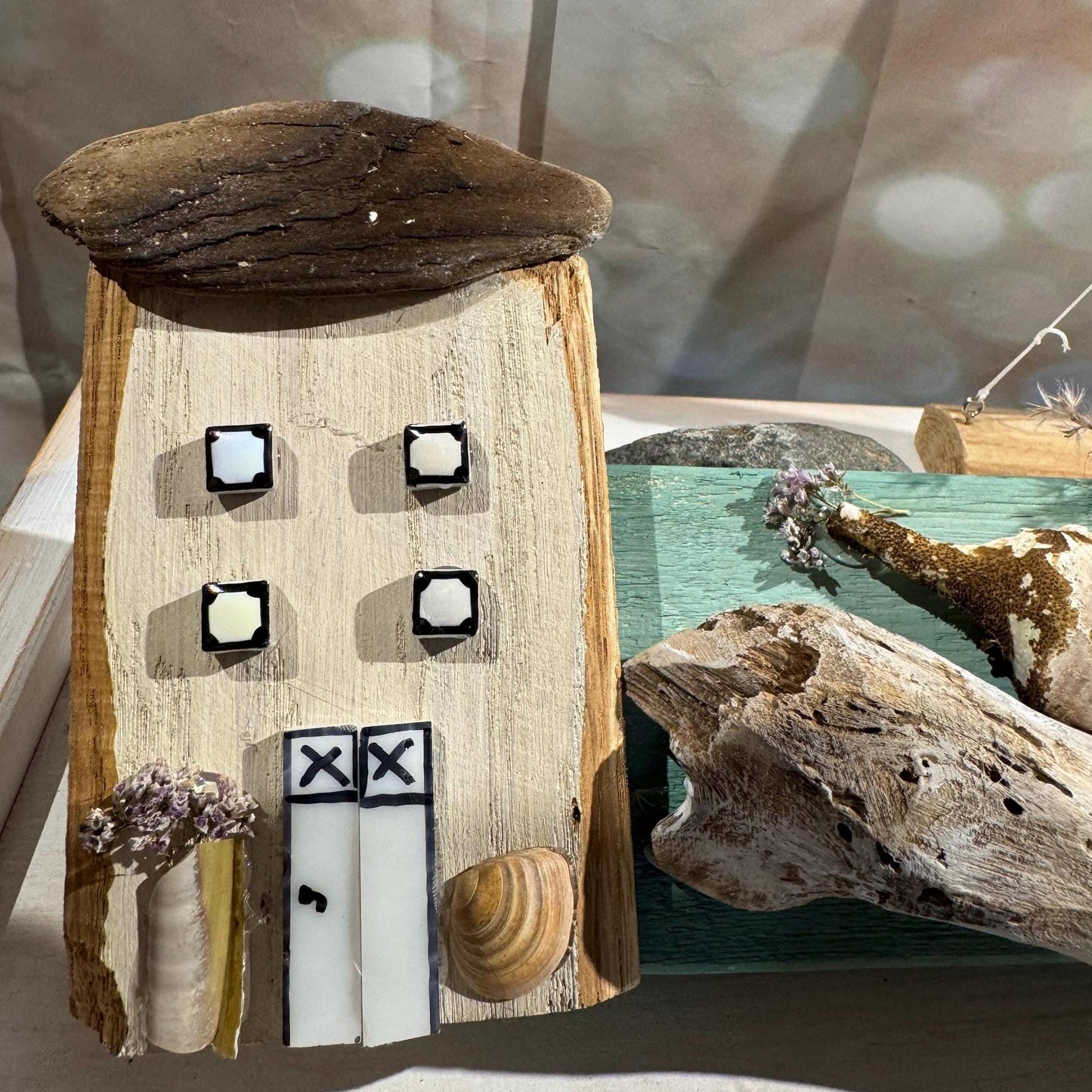Eine handgefertigte Holzhaus-Skulptur mit einem Treibholz-Dach, vier quadratischen Fenstern und zwei weißen Türen.
