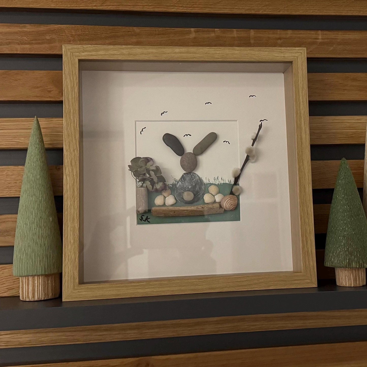 Gerahmtes Kunstwerk mit einem Hasen aus Kieselsteinen in Grau- und Brauntönen auf weißem Hintergrund.