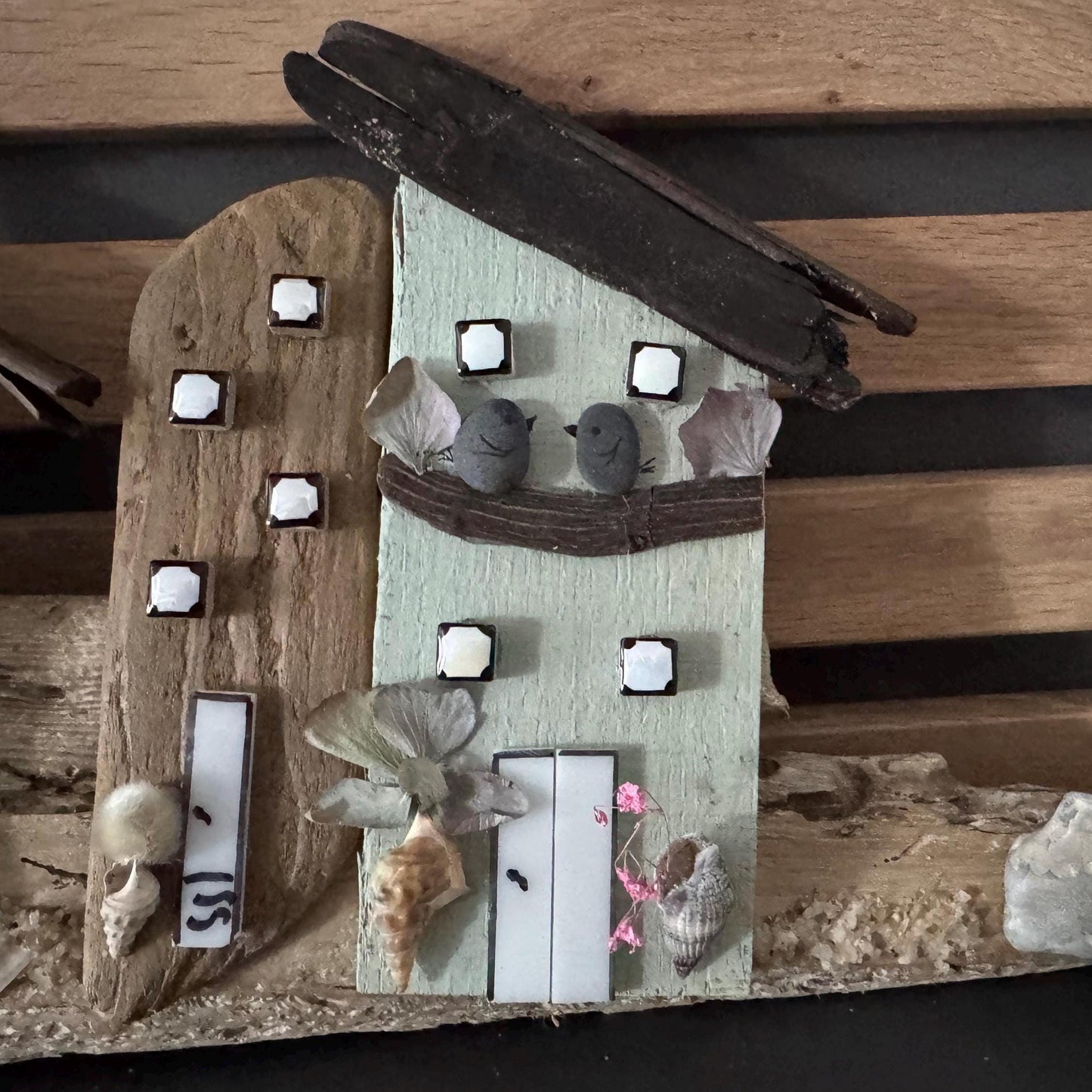Eine handgefertigte Holzhaus-Skulptur mit hellgrüner Fassade, quadratischen Fenstern und einem dunklen Holzdach.