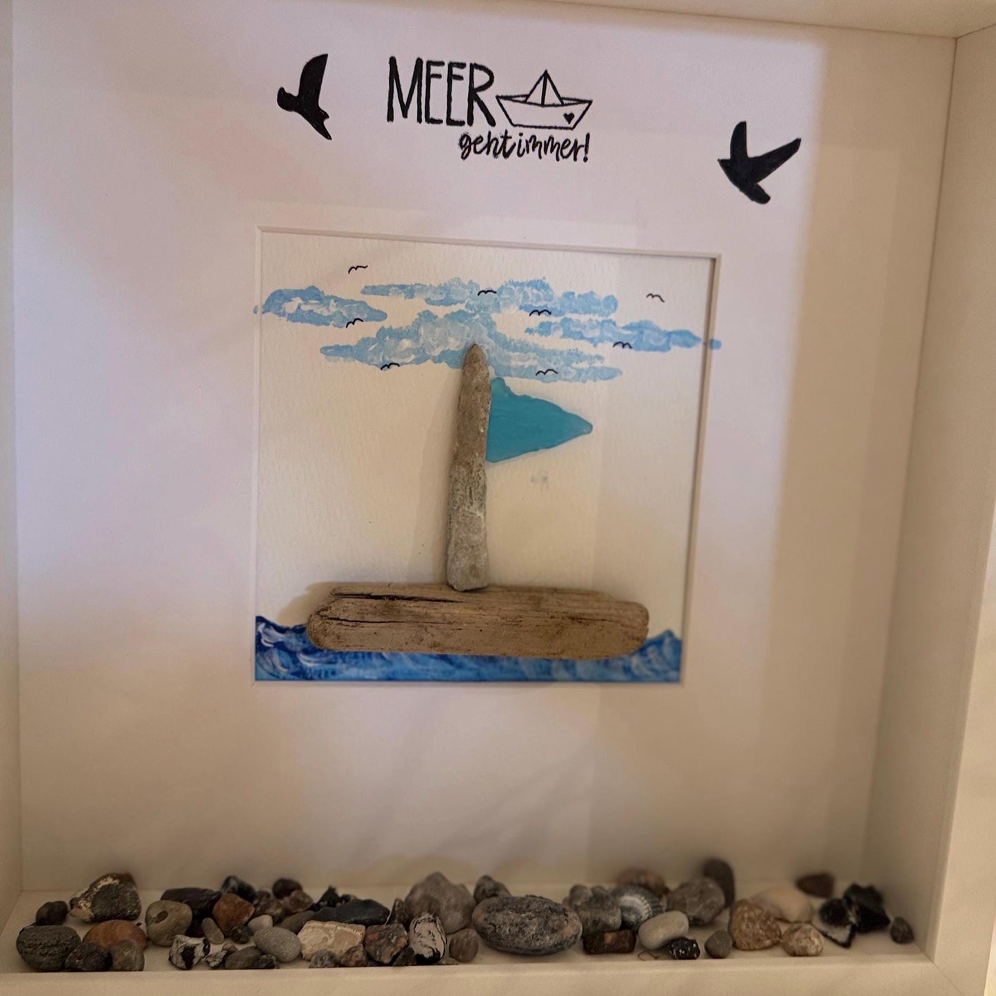Gerahmtes Kunstwerk mit einem Segelboot aus Treibholz und einem blauen Segel vor einem gemalten Himmel mit Wolken und Vögeln.