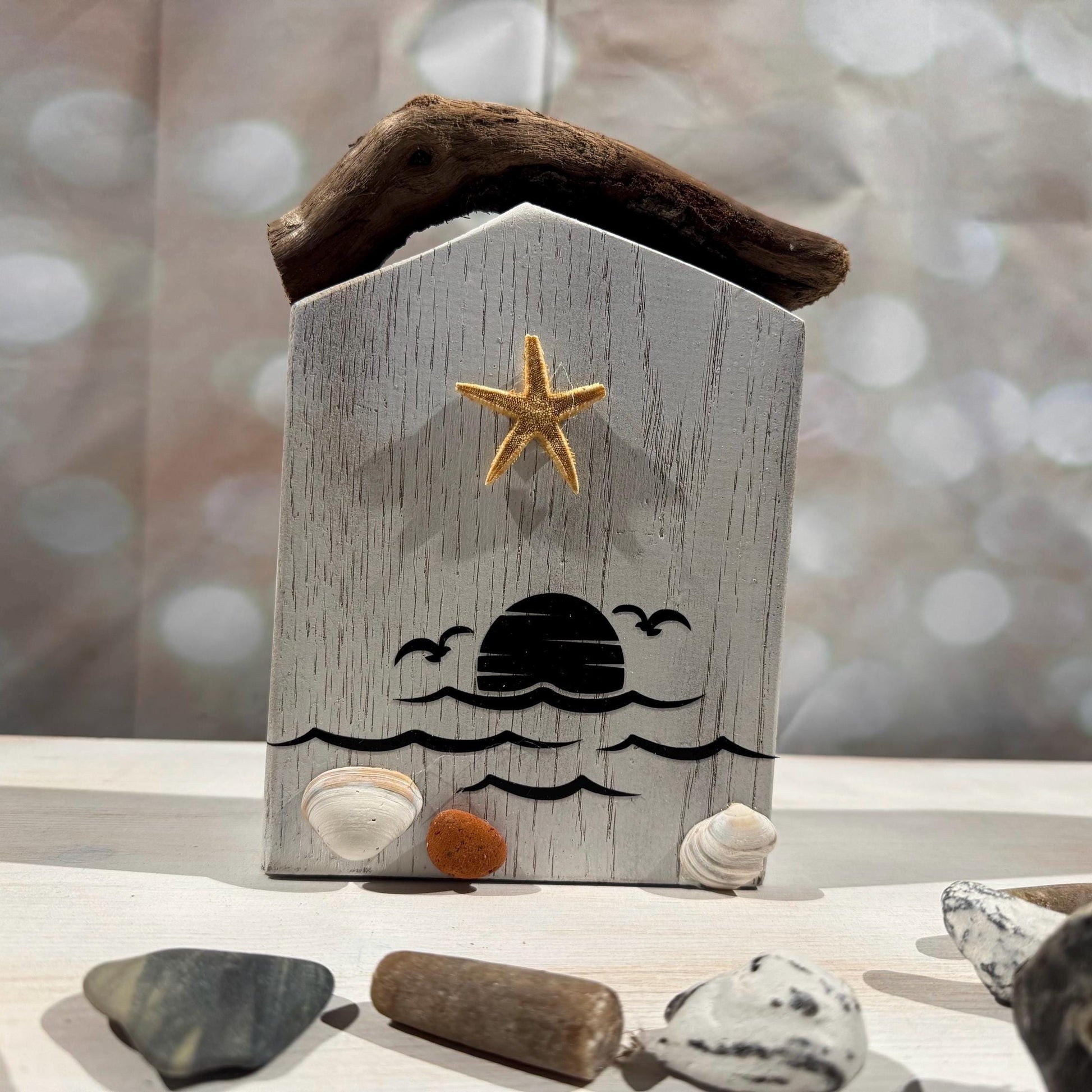 Ein dekoratives Strandhaus aus Holz mit einem Treibholz-Dach, einem goldenen Seestern und einem Sonnenuntergangs-Design.