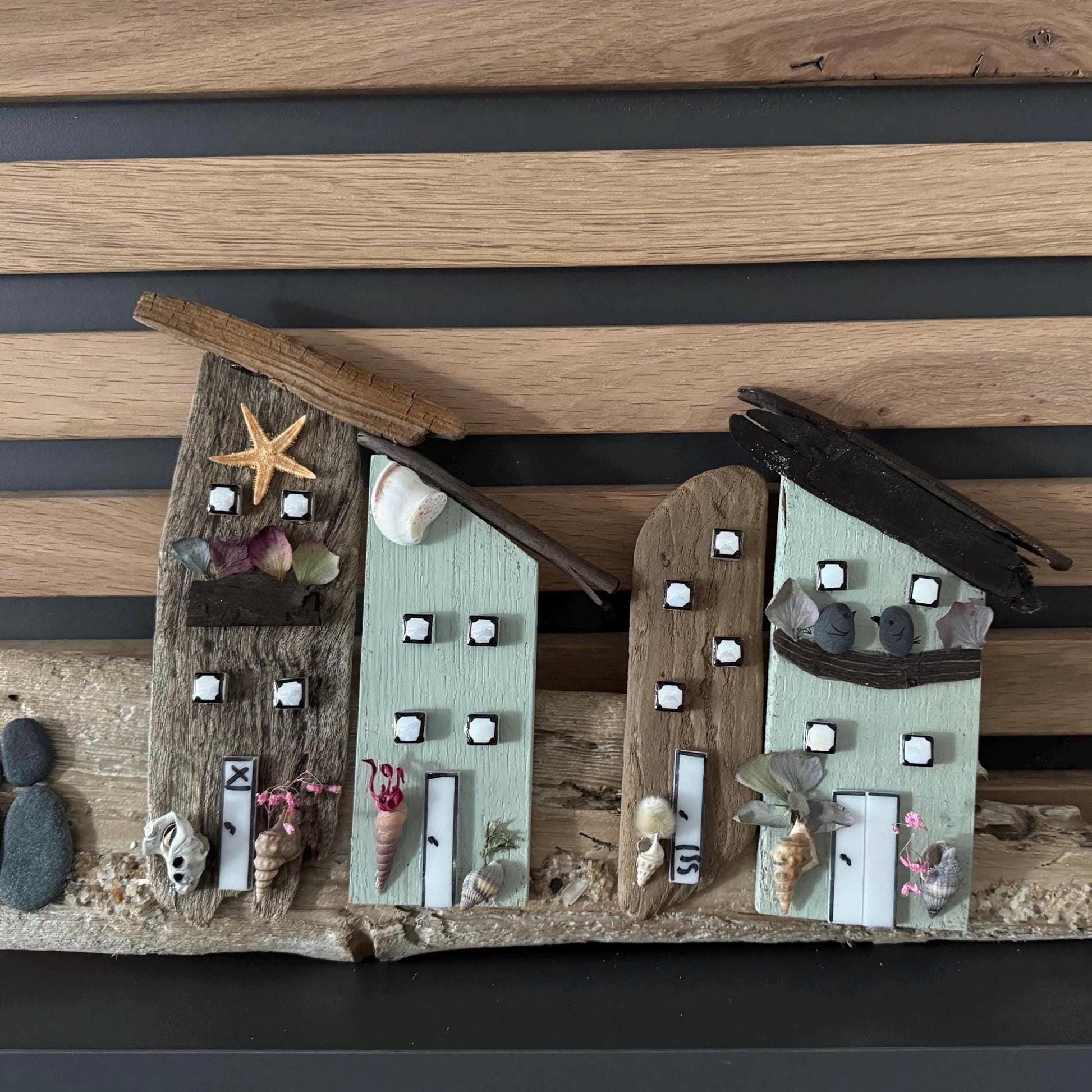 Künstlerisches Holzobjekt mit Küstenmotiv, das Miniaturhäuser aus Treibholz zeigt.  Das Kunstwerk ist auf einer Holzplanke