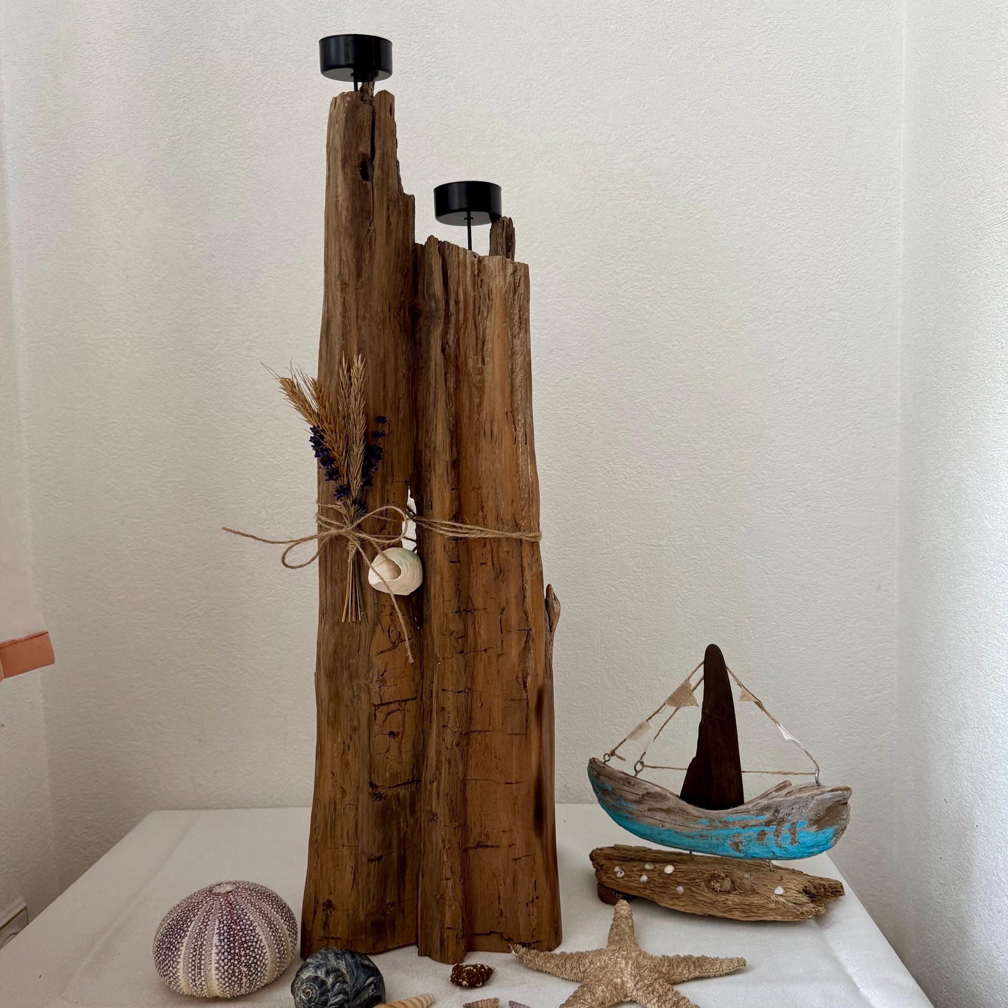 Ein rustikaler Kerzenhalter aus Holz mit zwei schwarzen Kerzenschalen, verziert mit Trockenblumen und einer Muschel.
