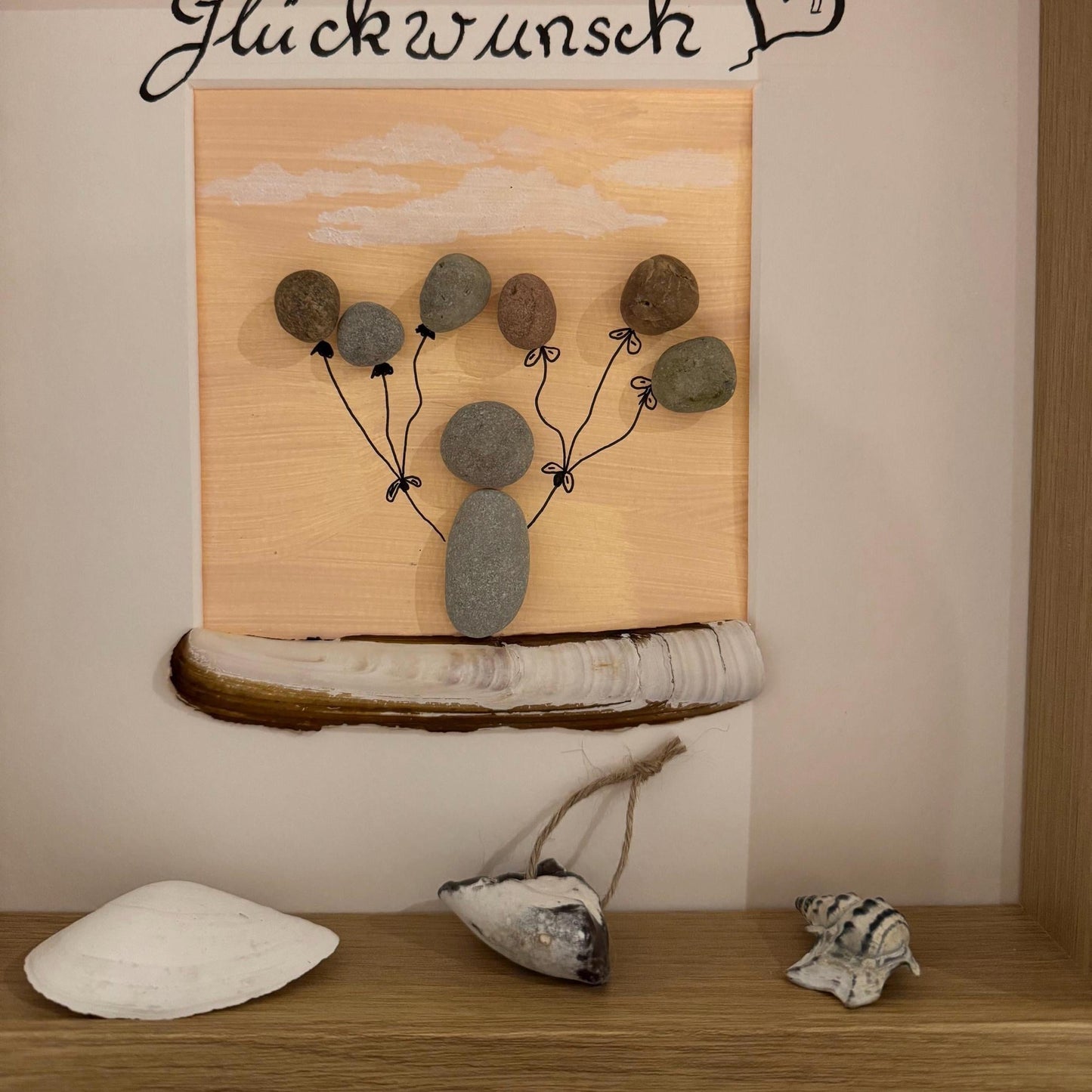 Gerahmtes Kunstwerk mit einer Kieselkunstfigur, die Kieselballons vor einem pfirsichfarbenen Hintergrund hält.