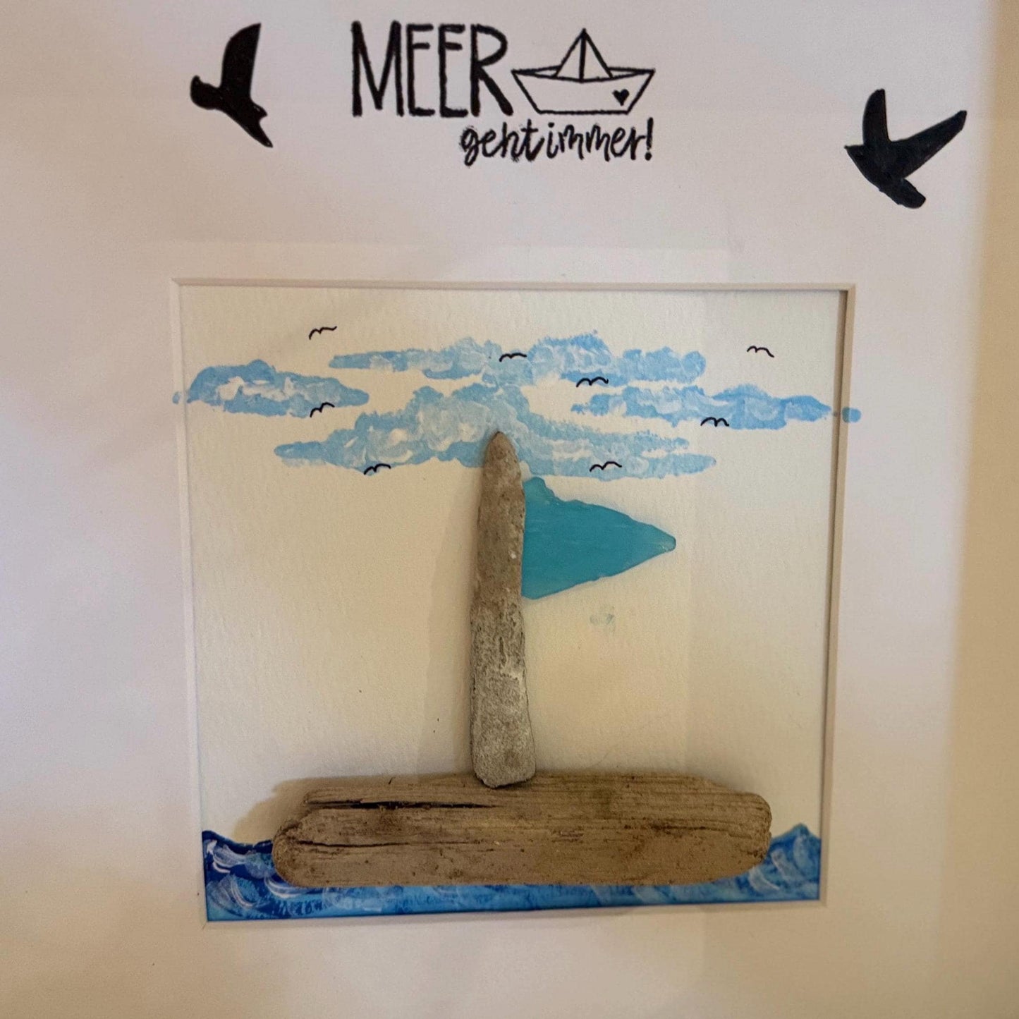 Gerahmtes Kunstwerk mit einem Segelboot aus Treibholz mit blauer Flagge, vor dem Hintergrund blauer Wolken und kleiner Vögel.