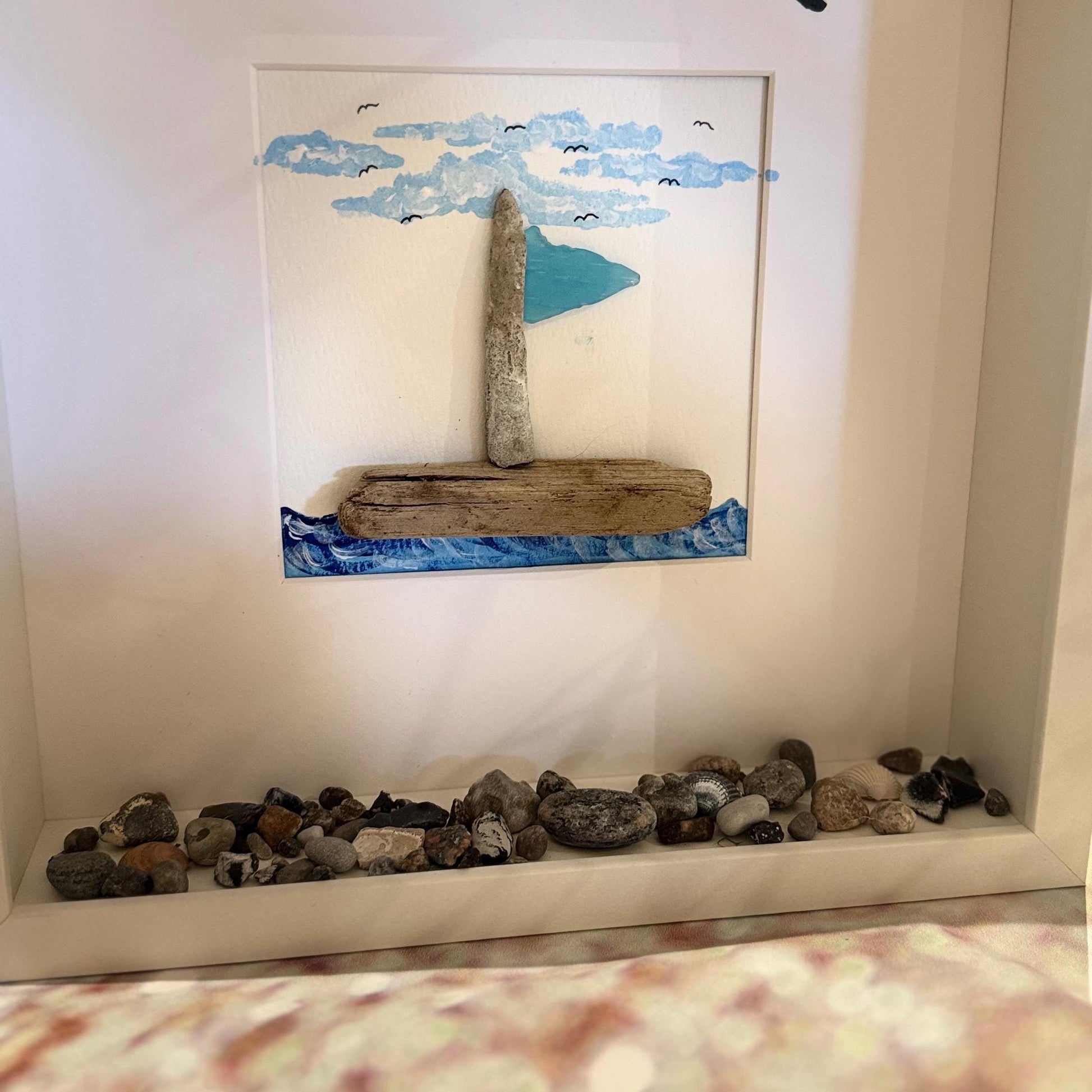 Ein gerahmtes nautisches Kunstwerk mit einem Segelboot aus Treibholz und Stein. Das Boot hat ein hellblaues Segel