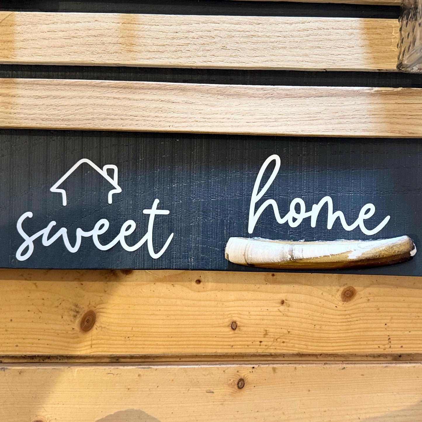 Ein Holzschild mit den Worten "sweet home" in weißer Schrift. Eine weiße Hausumrisslinie befindet sich über dem Wort "sweet".