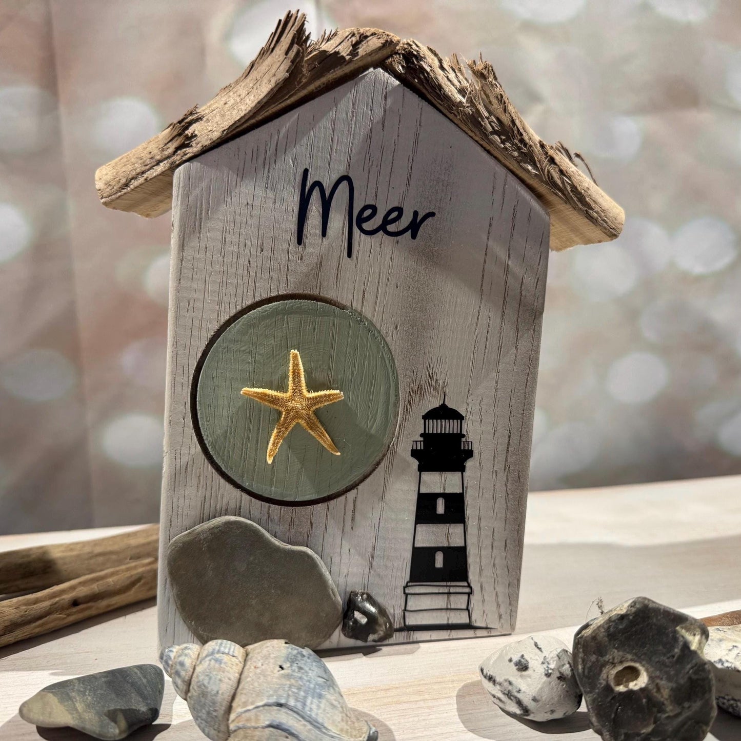Ein dekoratives Strandhaus aus Holz mit einem Treibholz-Dach. Das Haus ist hellgrau gestrichen und trägt den Schriftzug Meer