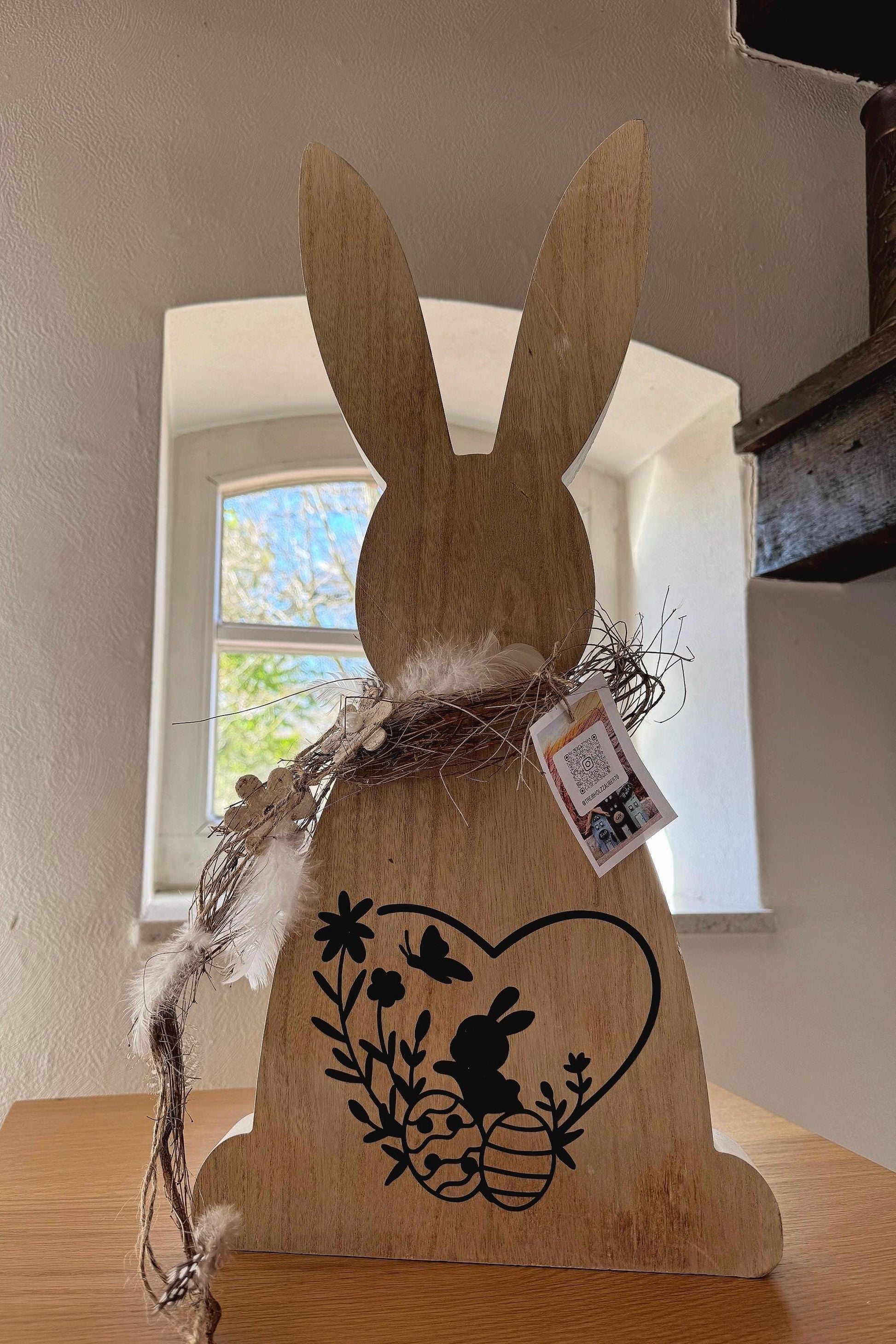 Holzdeko-Hase aus Naturholz mit schwarzem Oster-Motiv, verziert mit Federn und Kranz, handgemacht im rustikal-nordischen Stil