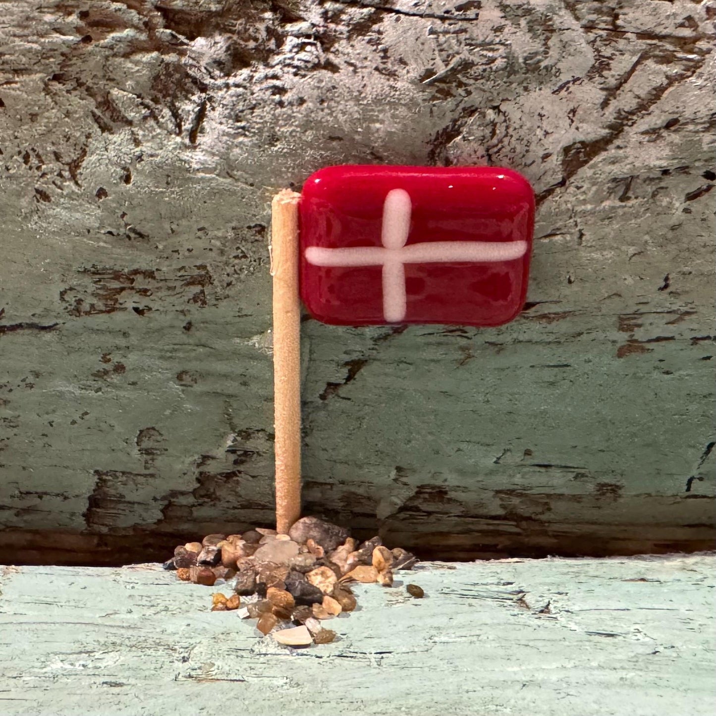 Eine kleine rote Flagge mit weißem Kreuz, befestigt an einem Holzstab, liegt auf einer Ansammlung winziger Kieselsteine.