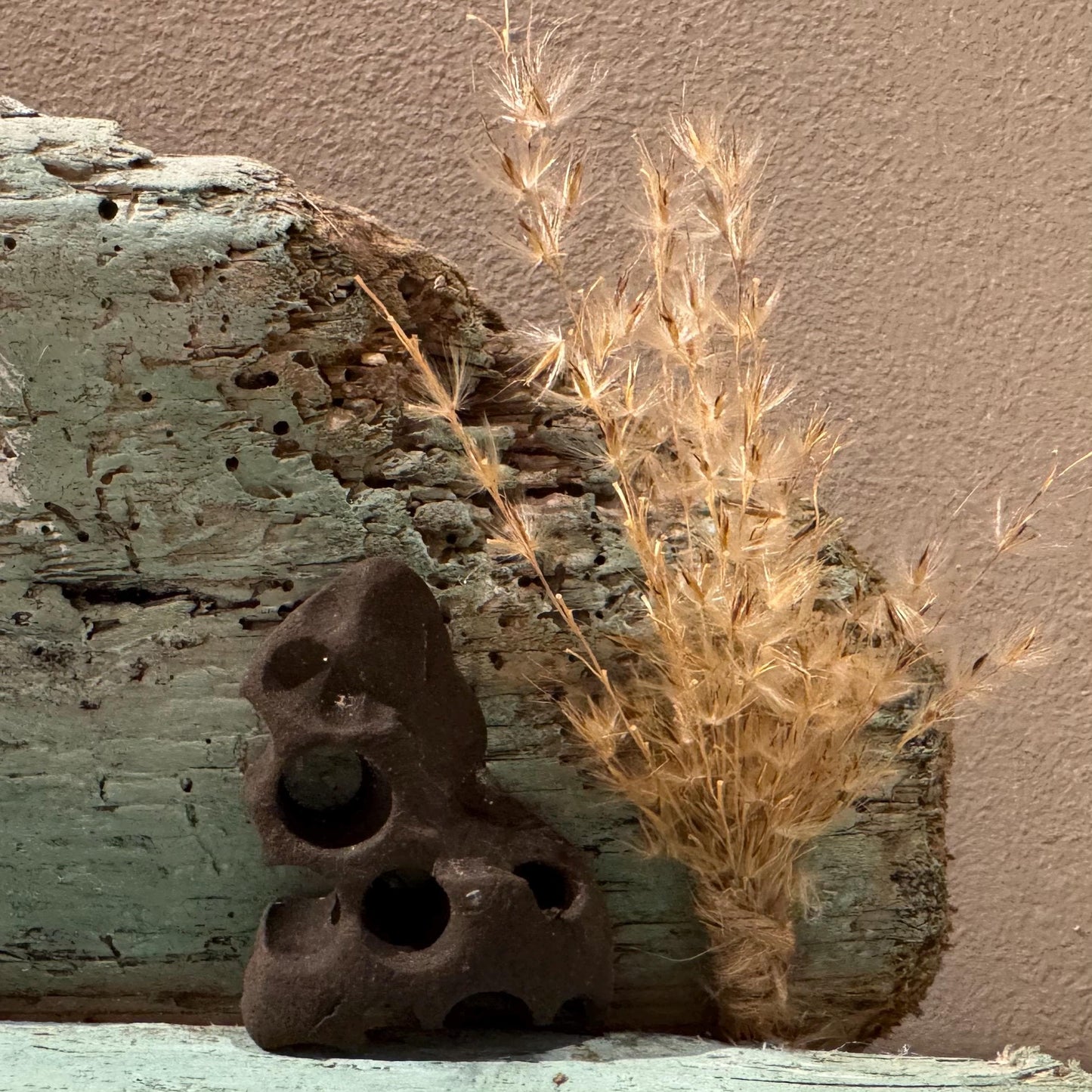 Eine dunkle, poröse, Steinskulptur mit mehreren kreisförmigen Öffnungen. Sie ruht auf einer verwitterten Holzoberfläche.