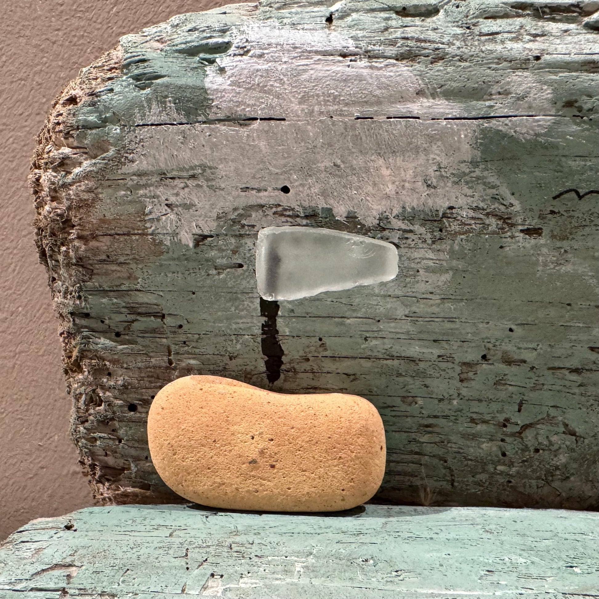 Ein Stück verwittertes, türkis lackiertes Holz mit einem hellen, rechteckigen Stück Meeresglas. Darunter liegt ein Stein.