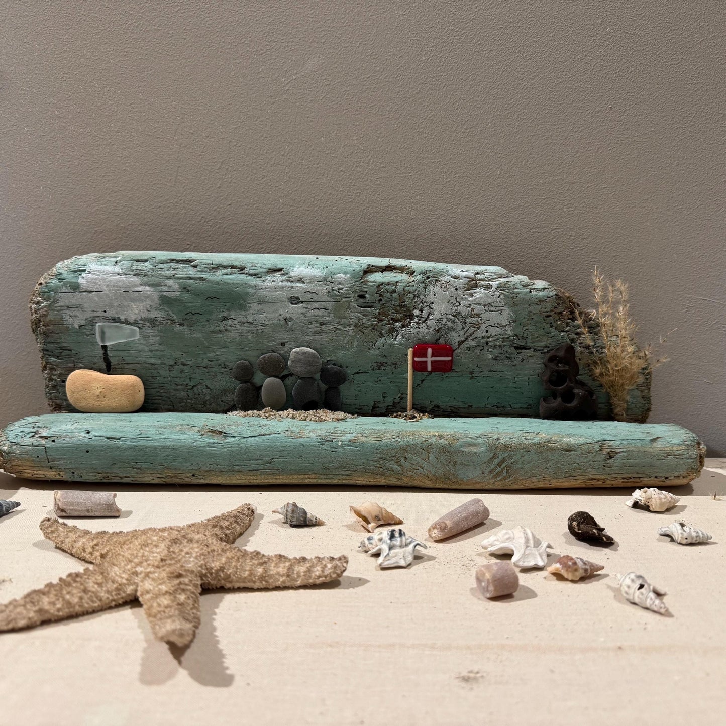 Ein Treibholz-Kunstwerk mit einer Strandszene mit einem Holzstamm und Kieselsteinfiguren was ein Küstenmotiv ergibt.