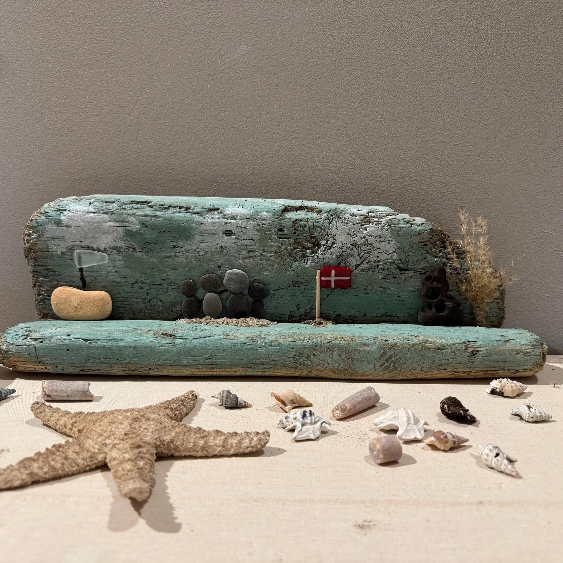 Ein Treibholz-Kunstwerk mit einer Strandszene mit einem Holzstamm und Kieselsteinfiguren was ein Küstenmotiv ergibt.