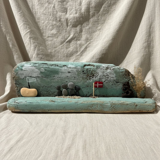 Ein Treibholz-Kunstwerk mit einer Strandszene mit einem Holzstamm und Kieselsteinfiguren was ein Küstenmotiv ergibt.