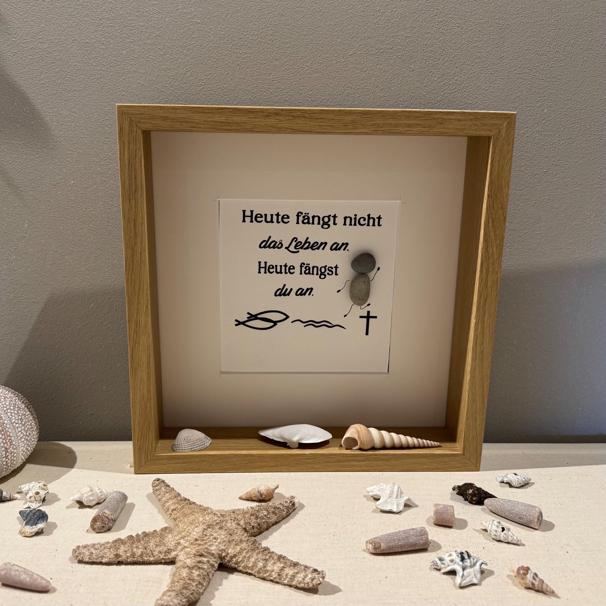 Ein gerahmtes Kunstwerk mit einem hellen Holzrahmen. Das Kunstwerk zeigt einen weißen Hintergrund mit Text und Kieselkunst.