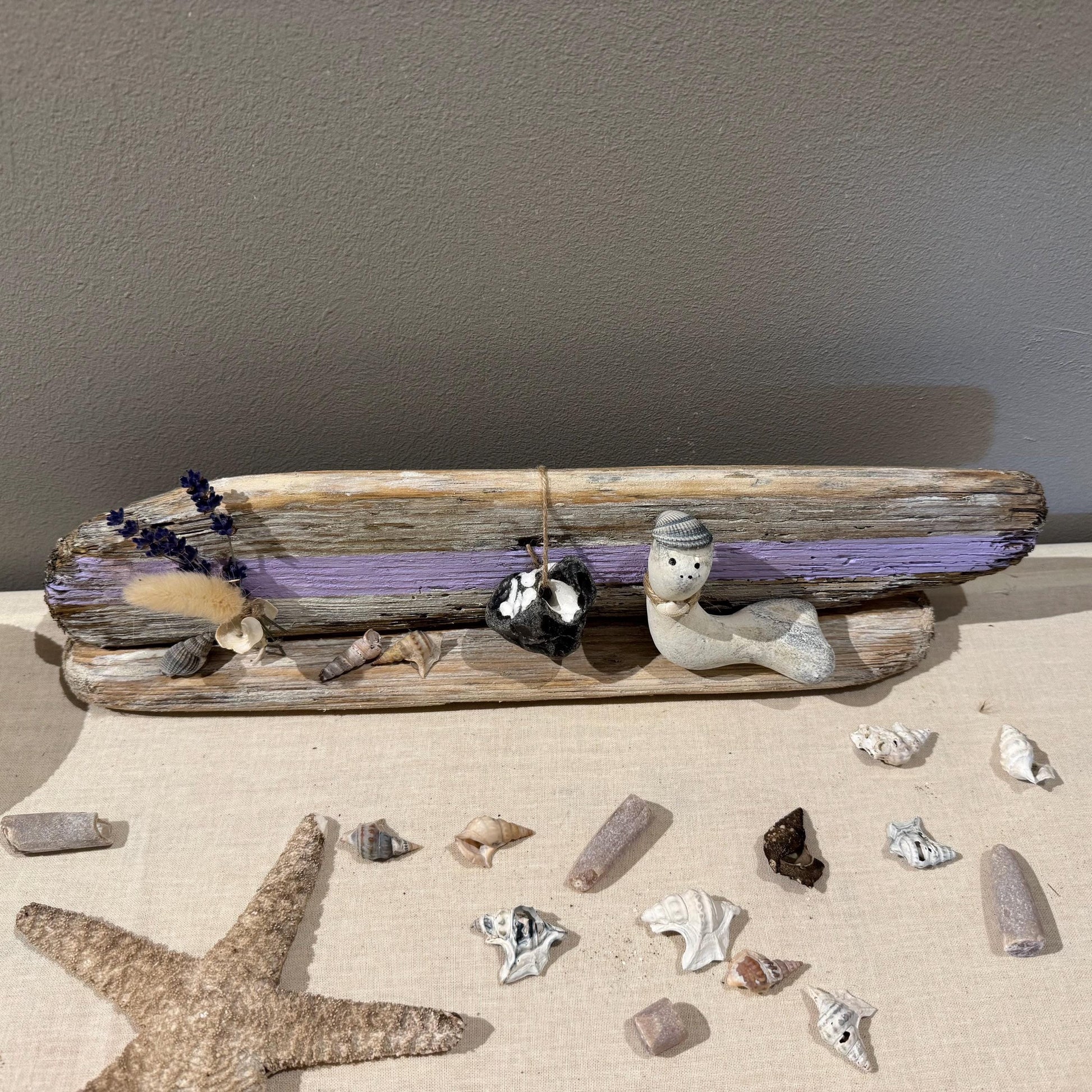 Maritime Treibholz Deko mit kleiner Steinfigur und Muscheln, liebevoll arrangiert auf Treibholz vor einer gemütlichen Couch.