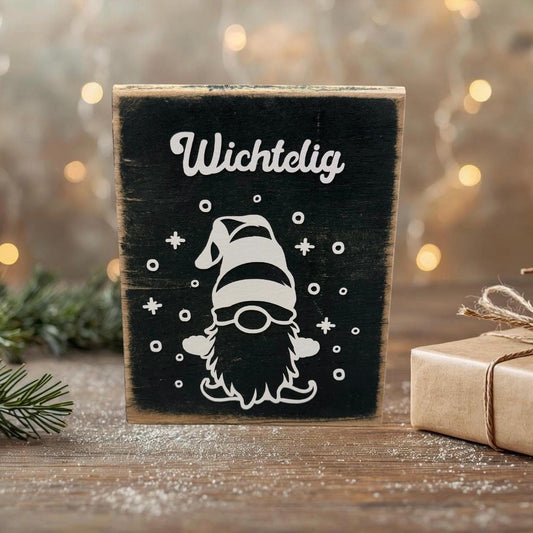 Rustikale Handgefertigte Deko mit Weihnachtswichtel mit weißem „Wichtelig“-Motiv auf schwarzer Tafel, winterlich inszeniert mit Tannenzweig, Geschenk und funkelnden Lichtern im Hintergrund.