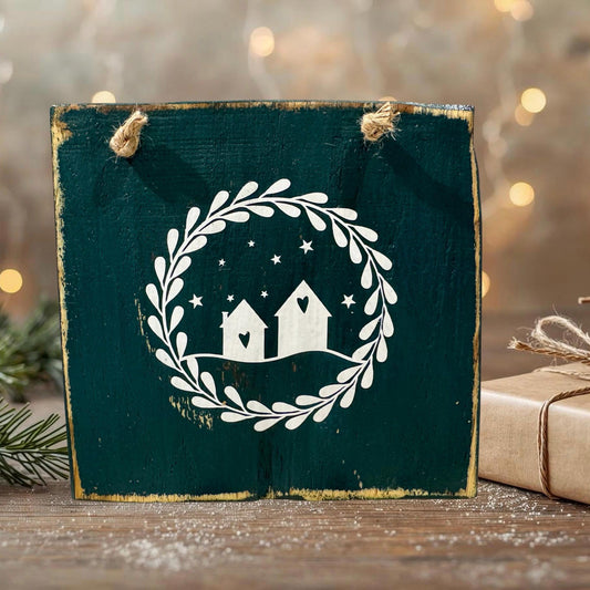 Rustikale grüne Holz-Deko mit weißem Häuserkranz-Motiv, aufgehängt an Juteseilen, auf einem Holztisch mit weihnachtlicher Hintergrundbeleuchtung.