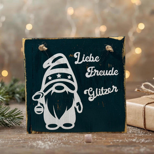 Holzschild im rustikalen Look mit weißer Wichtel-Silhouette und dem Schriftzug „Liebe Freude Glitzer“, dekoriert mit einer Juteschnur zum Aufhängen. Im Hintergrund liegen Tannenzweige, ein Geschenk und warme Lichter für eine gemütliche Weihnachtsstimmung.