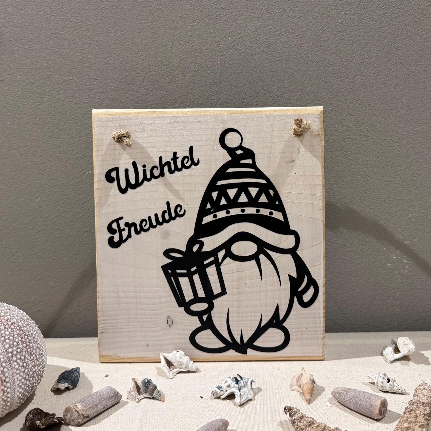 Ein helles Holzschild mit dem Schriftzug "Wichtel Freude" und einer schwarzen Zwerg-Illustration, die ein Geschenk hält.