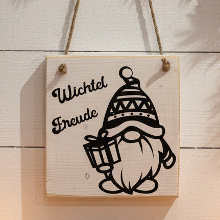 Quadratisches Holzschild mit dem Schriftzug "Wichtel Freude" und einer schwarzen Illustration eines Wichtels mit Geschenk.