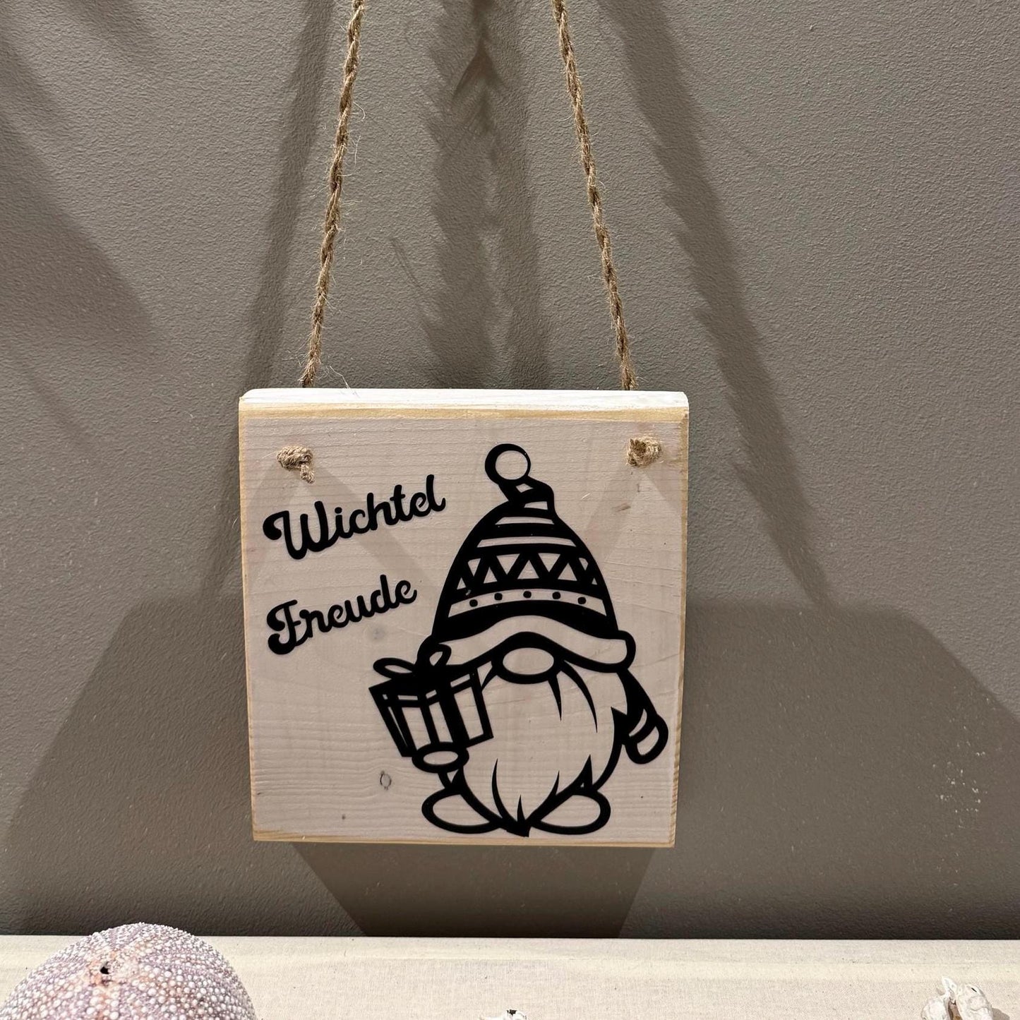 Quadratisches Holzschild mit dem Schriftzug "Wichtel Freude" und einer schwarzen Illustration eines Wichtels mit Geschenk.