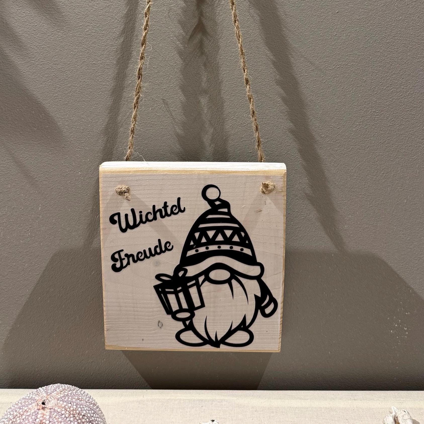 Quadratisches Holzschild mit dem Schriftzug "Wichtel Freude" und einer schwarzen Illustration eines Wichtels mit Geschenk.