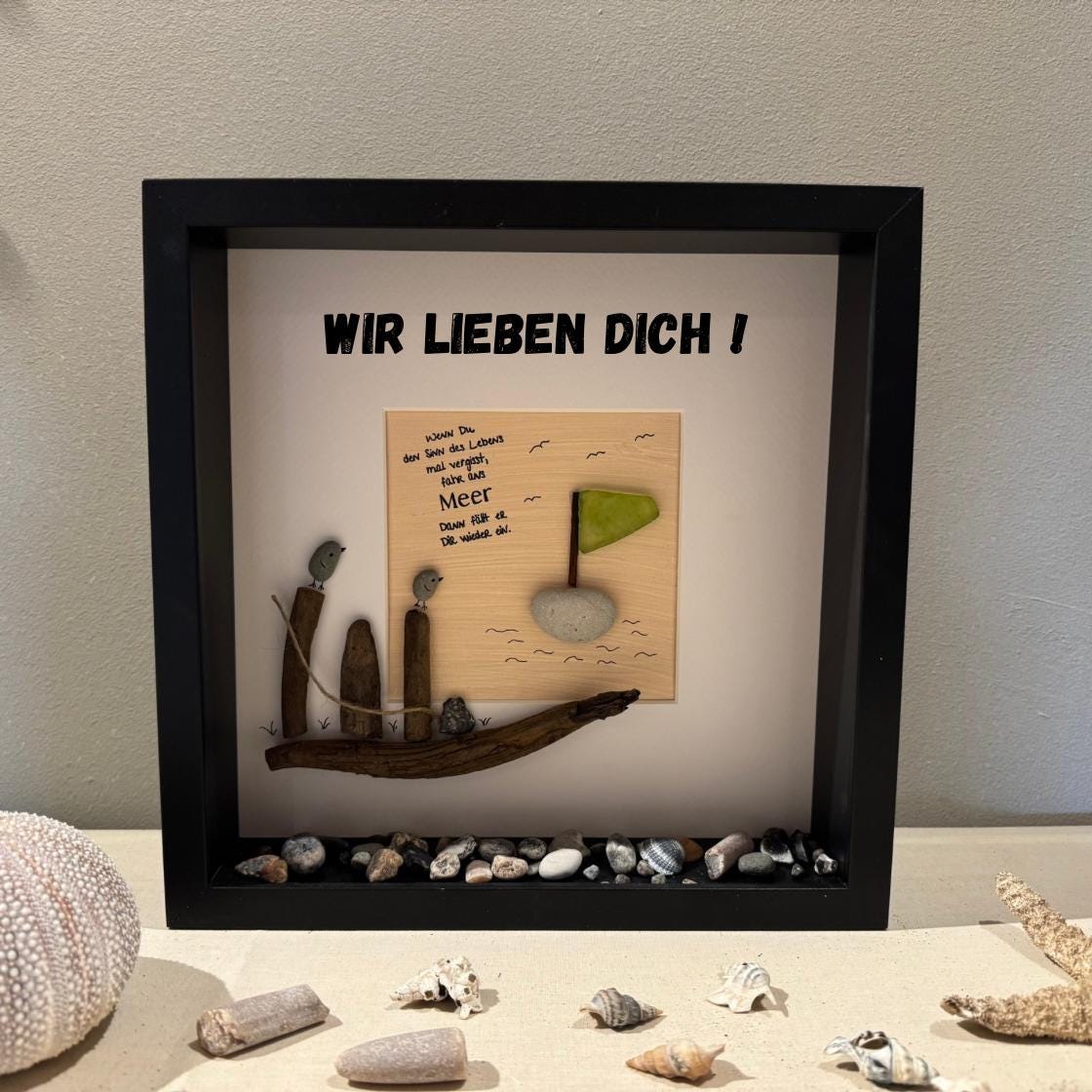 Ein gerahmtes Kunstwerk mit schwarzem Rahmen. Das Kunstwerk zeigt ein Treibholzboot mit zwei Vogelfiguren