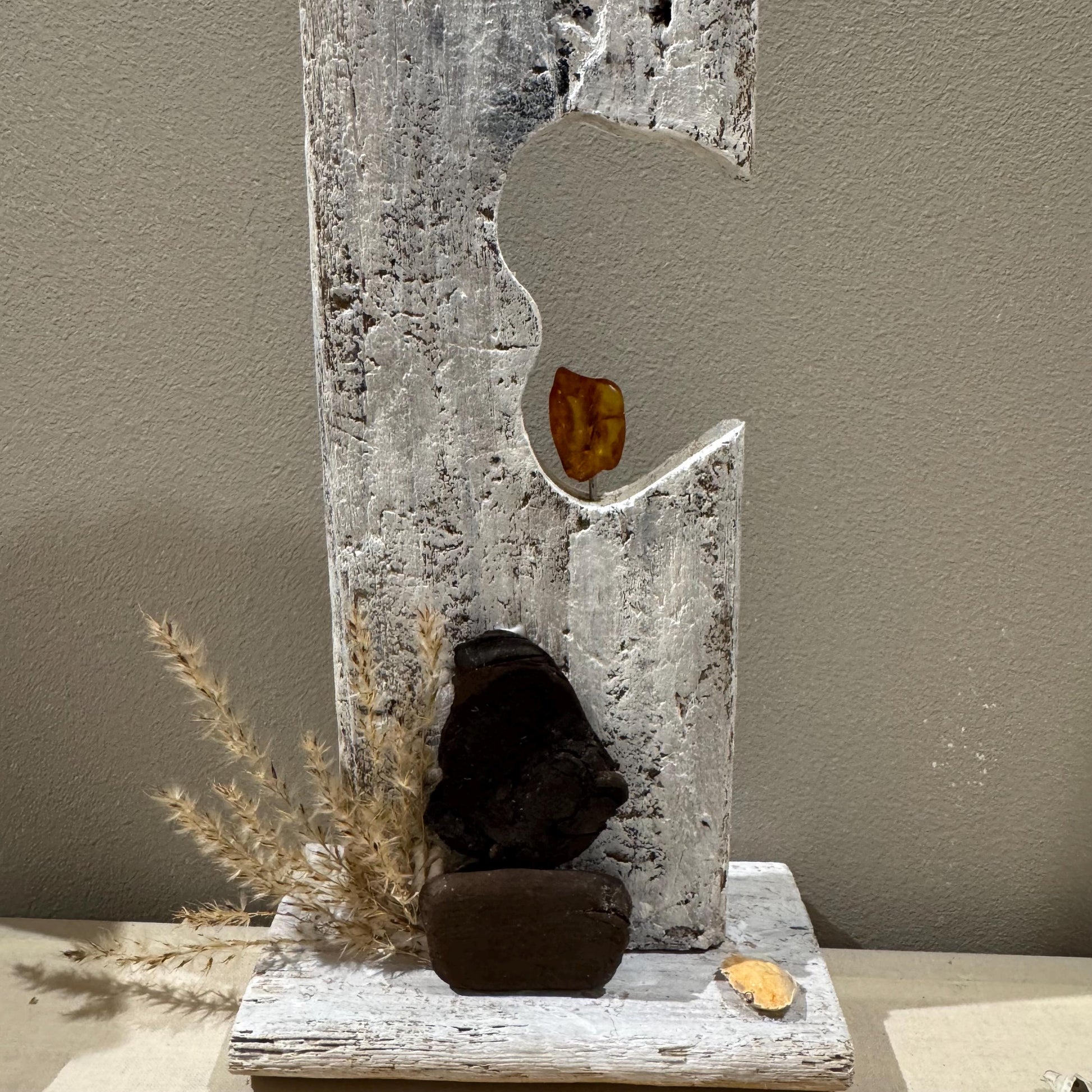 Rustikale, abstrakte Holzskulptur mit verwitterter, weiß lackierter Oberfläche. Getrocknete Gräser ergänzen das Design.