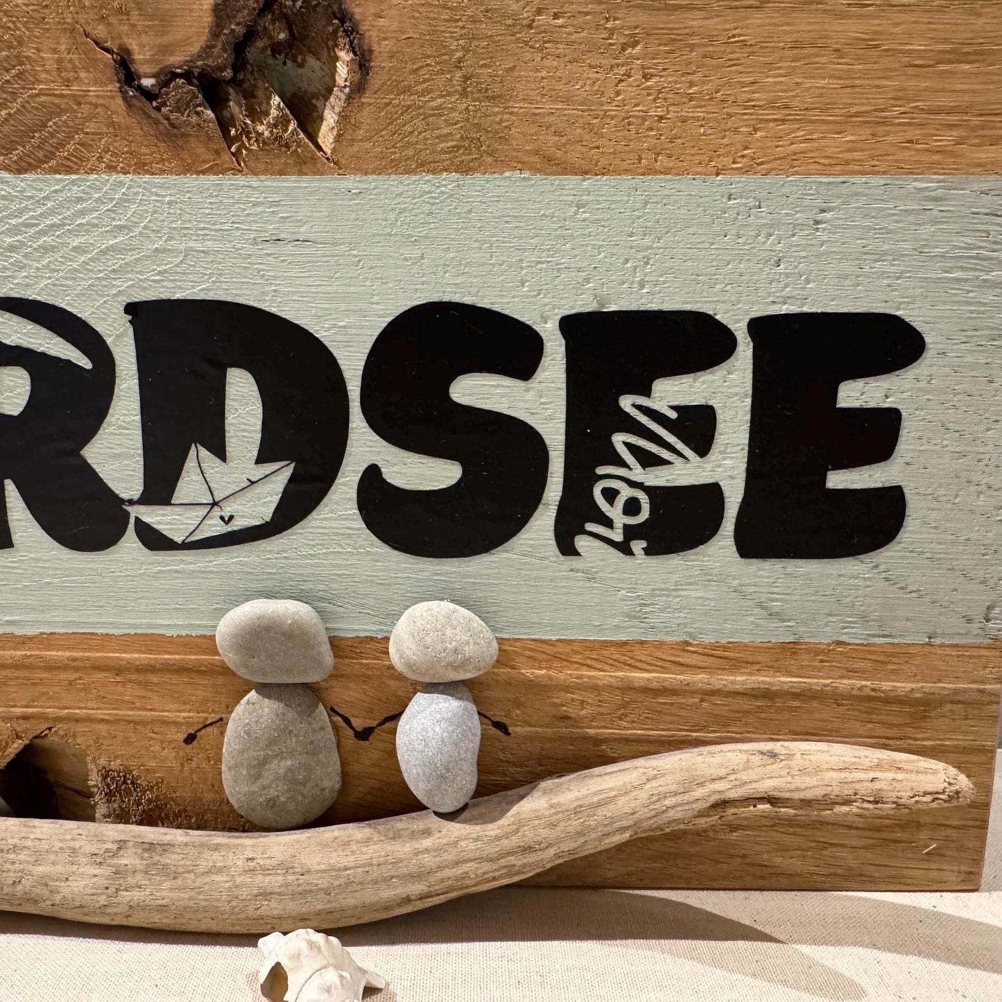 Holzschild mit dem Wort "NORDSEE" in Schwarz, mit einer Segelbootgrafik. Zwei Kieselsteinfiguren halten sich an den Händen.