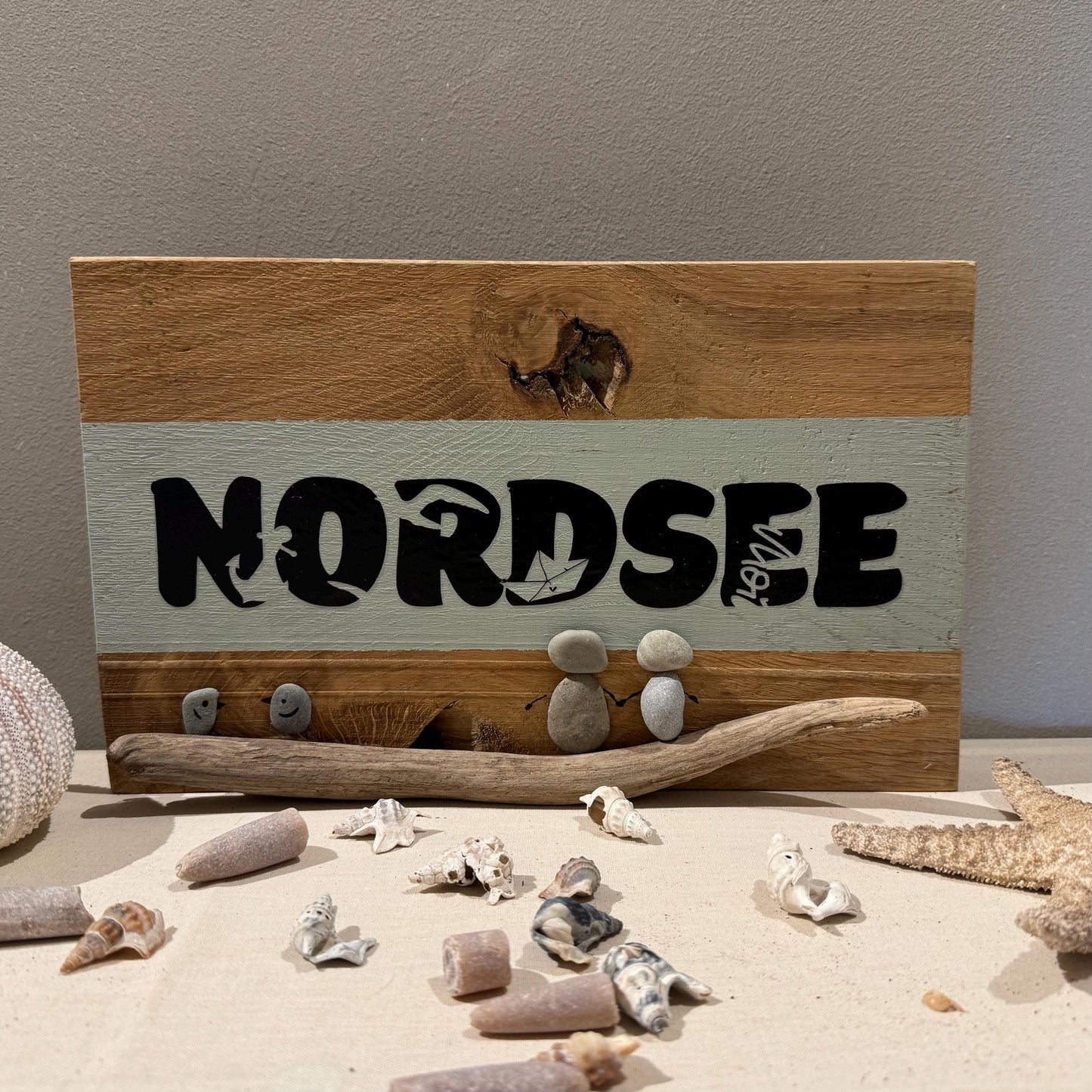 Holzschild mit dem Wort "NORDSEE" in Schwarz. Das Schild hat einen hellgrünen Streifen und zeigt Kieselkunstfiguren.