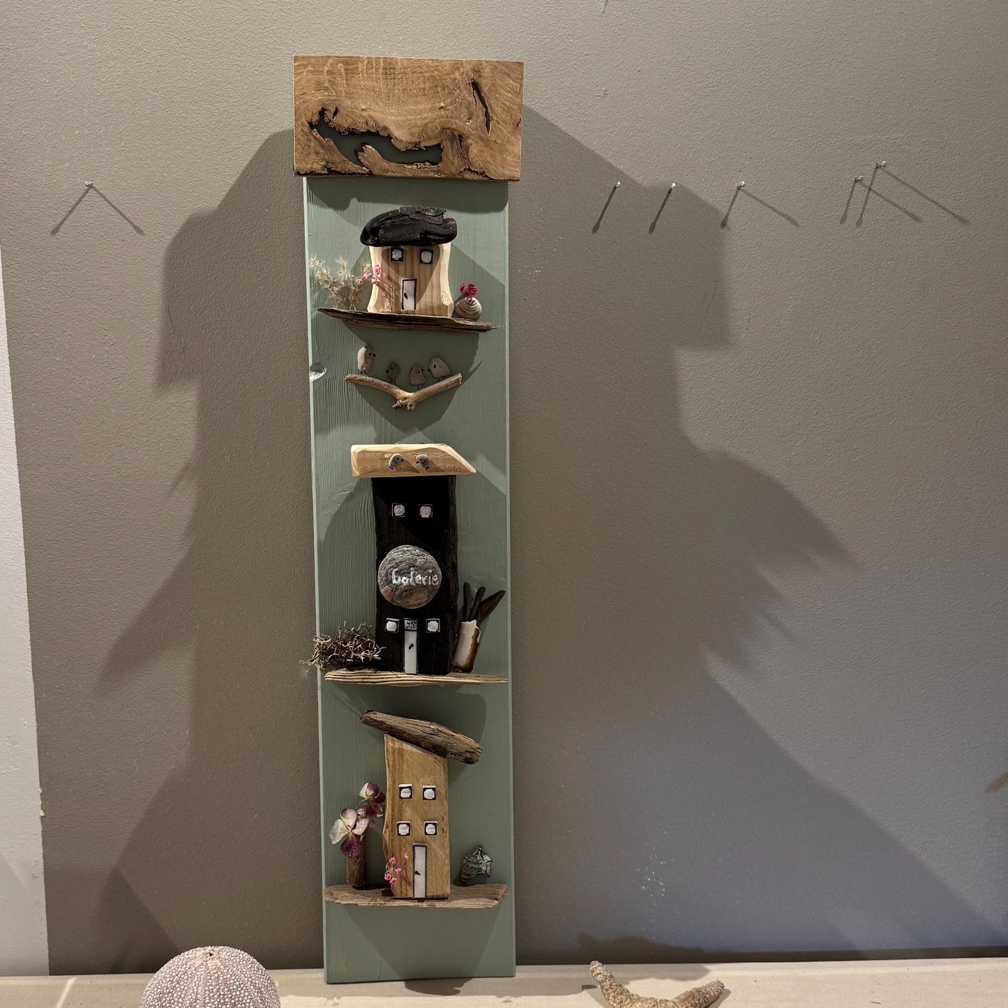 Ein vertikales Wandkunstwerk aus Holz mit Miniaturhäusern und Regalen. Die Häuser sind in verschiedenen Farben bemalt.