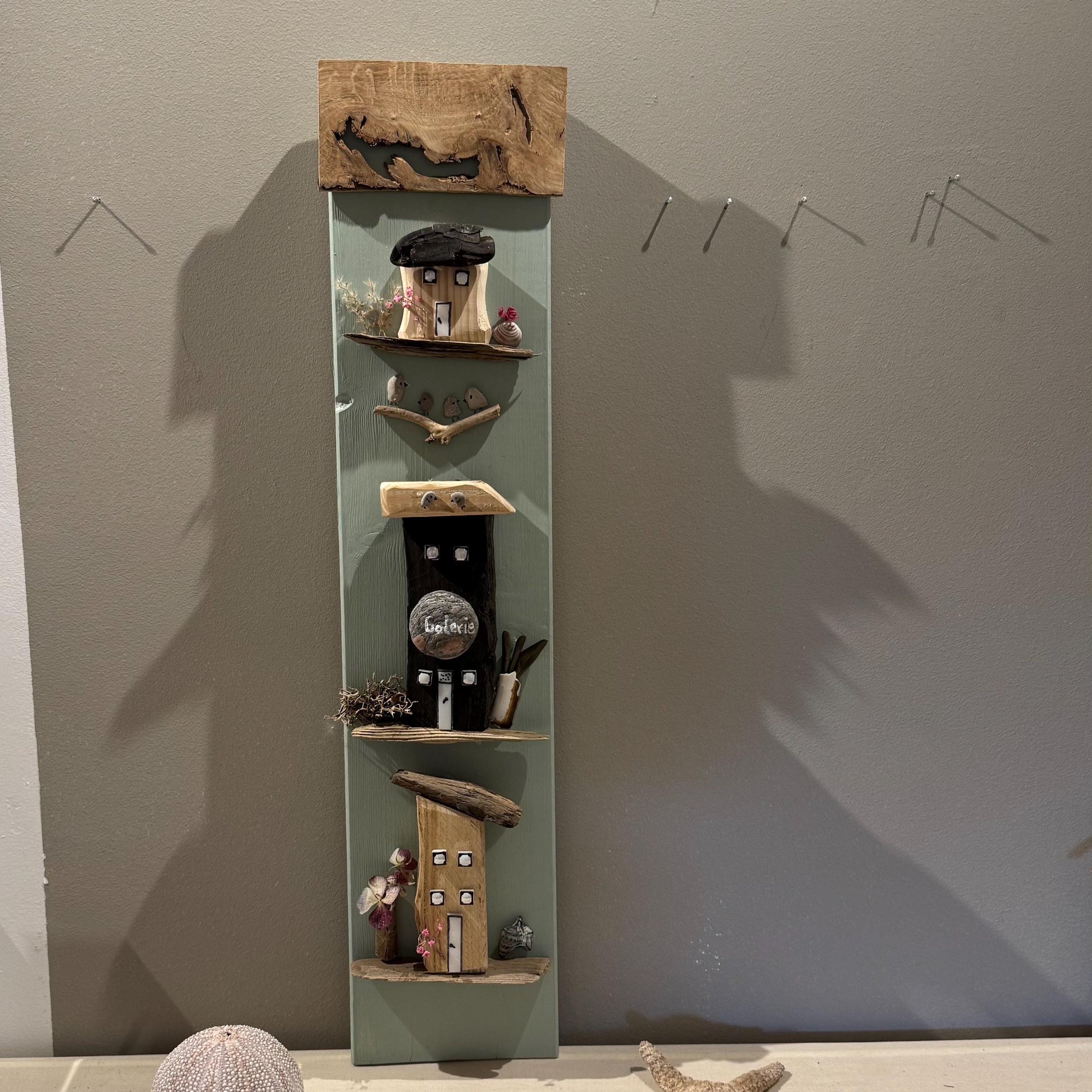 Ein vertikales Wandkunstwerk aus Holz mit Miniaturhäusern und Regalen. Die Häuser sind in verschiedenen Farben bemalt.