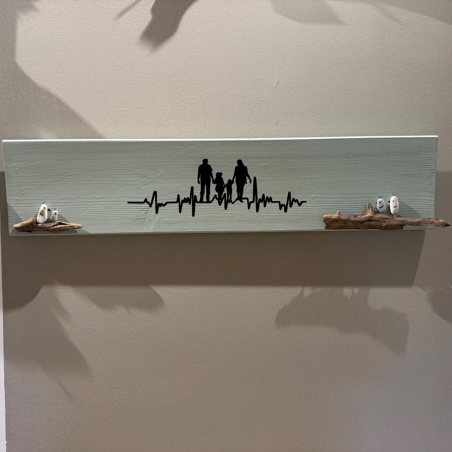 Ein hellgrünes Holzregal mit einer schwarzen Familiensilhouette und einem Herzschlag-Design von Treibholzzauber79