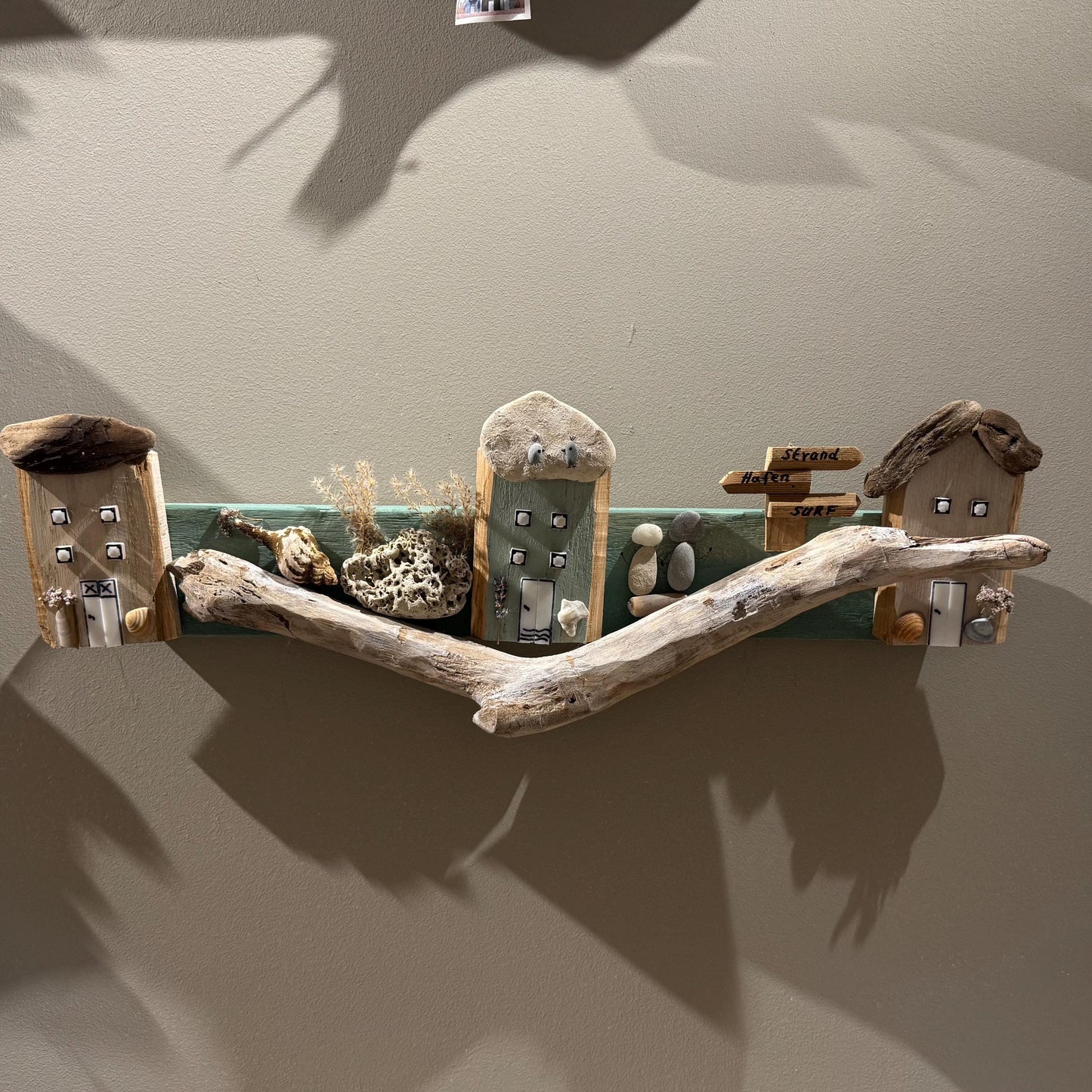 Ein Wandkunstwerk aus Treibholz mit Miniatur-Holzhäusern, einem Wegweiser und natürlichen Elementen.