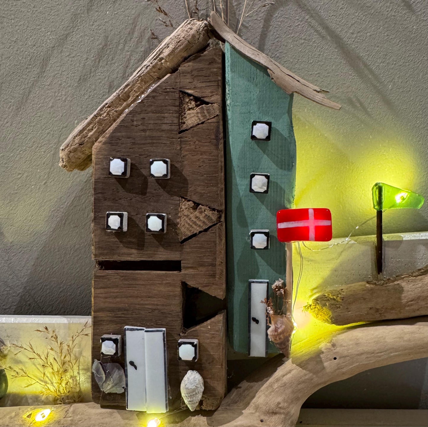 Treibholz Wanddeko Haus aus Treibholz mit kleinen Minihäusern und warmen LED-Lichtern auf heller Wand, maritime Dekoration.