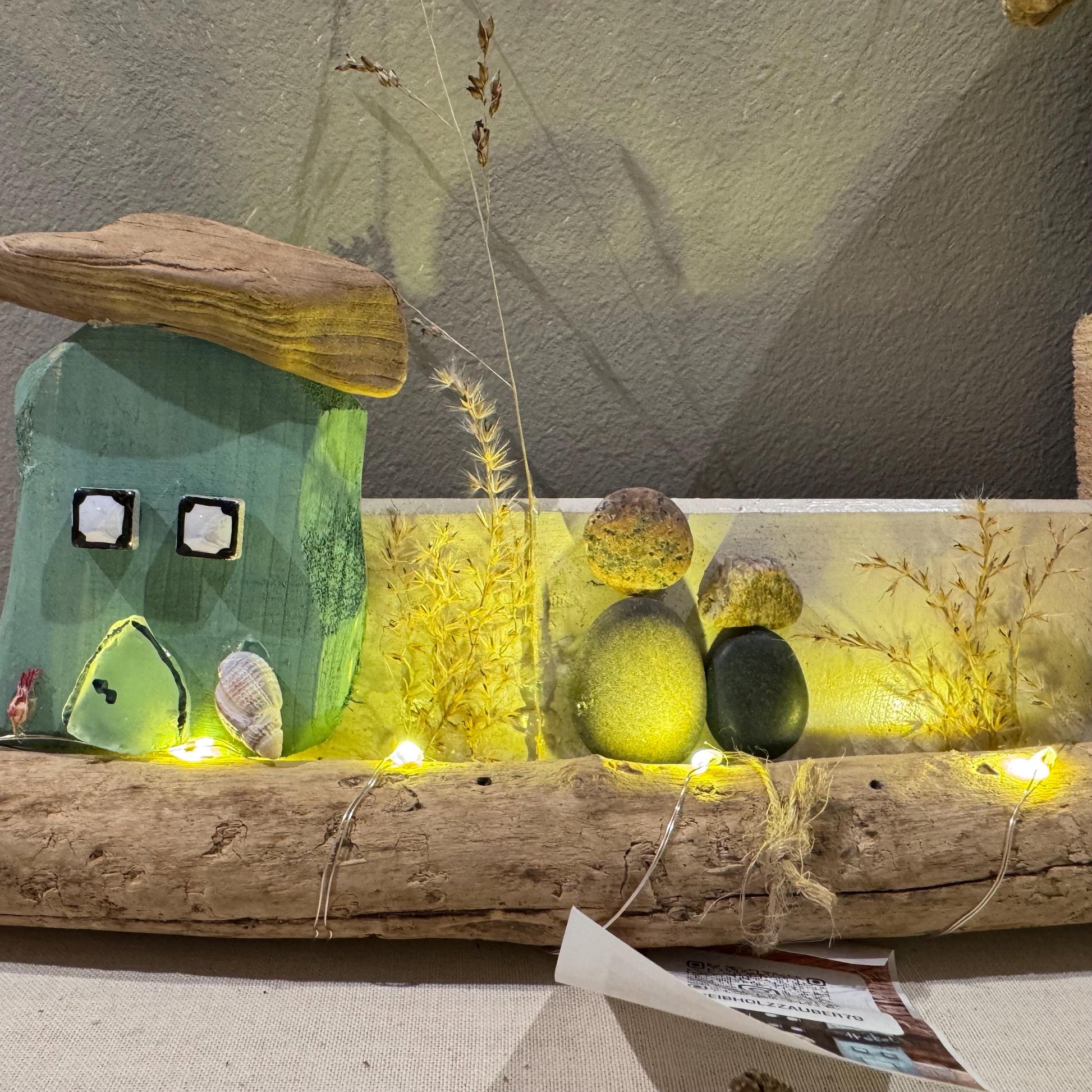 Treibholz Wanddeko Haus aus Treibholz mit kleinen Minihäusern und warmen LED-Lichtern auf heller Wand, maritime Dekoration.
