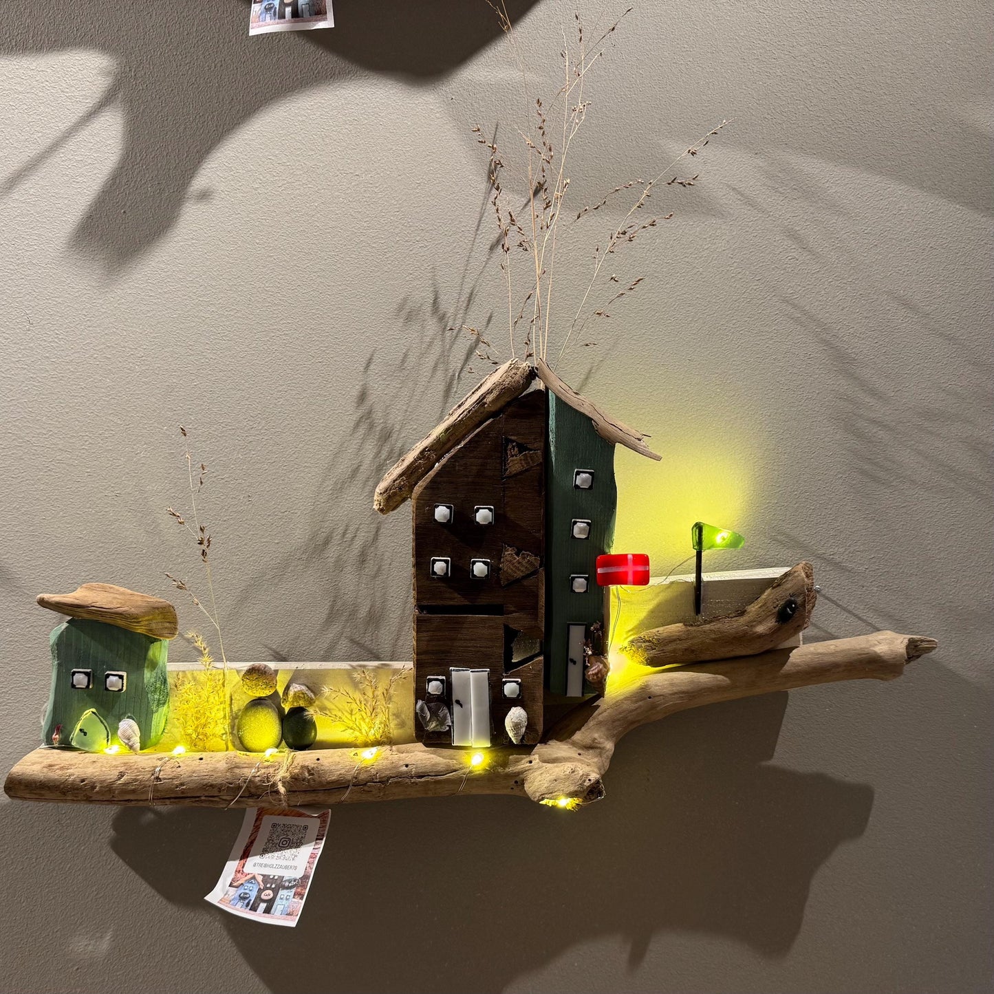 Treibholz Wanddeko Haus aus Treibholz mit kleinen Minihäusern und warmen LED-Lichtern auf heller Wand, maritime Dekoration.