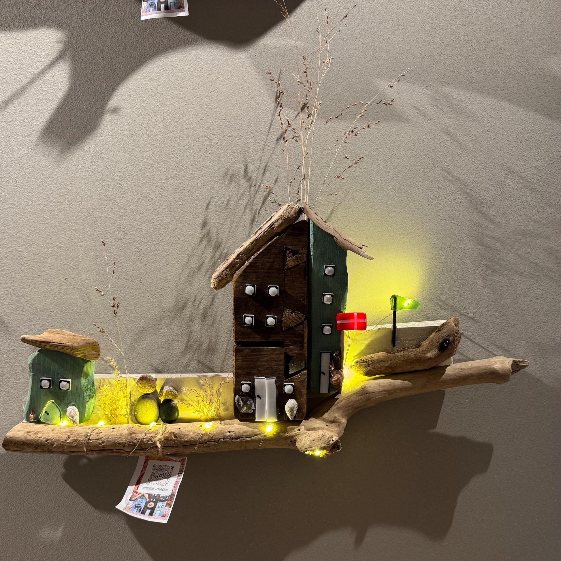 Treibholz Wanddeko Haus aus Treibholz mit kleinen Minihäusern und warmen LED-Lichtern auf heller Wand, maritime Dekoration.