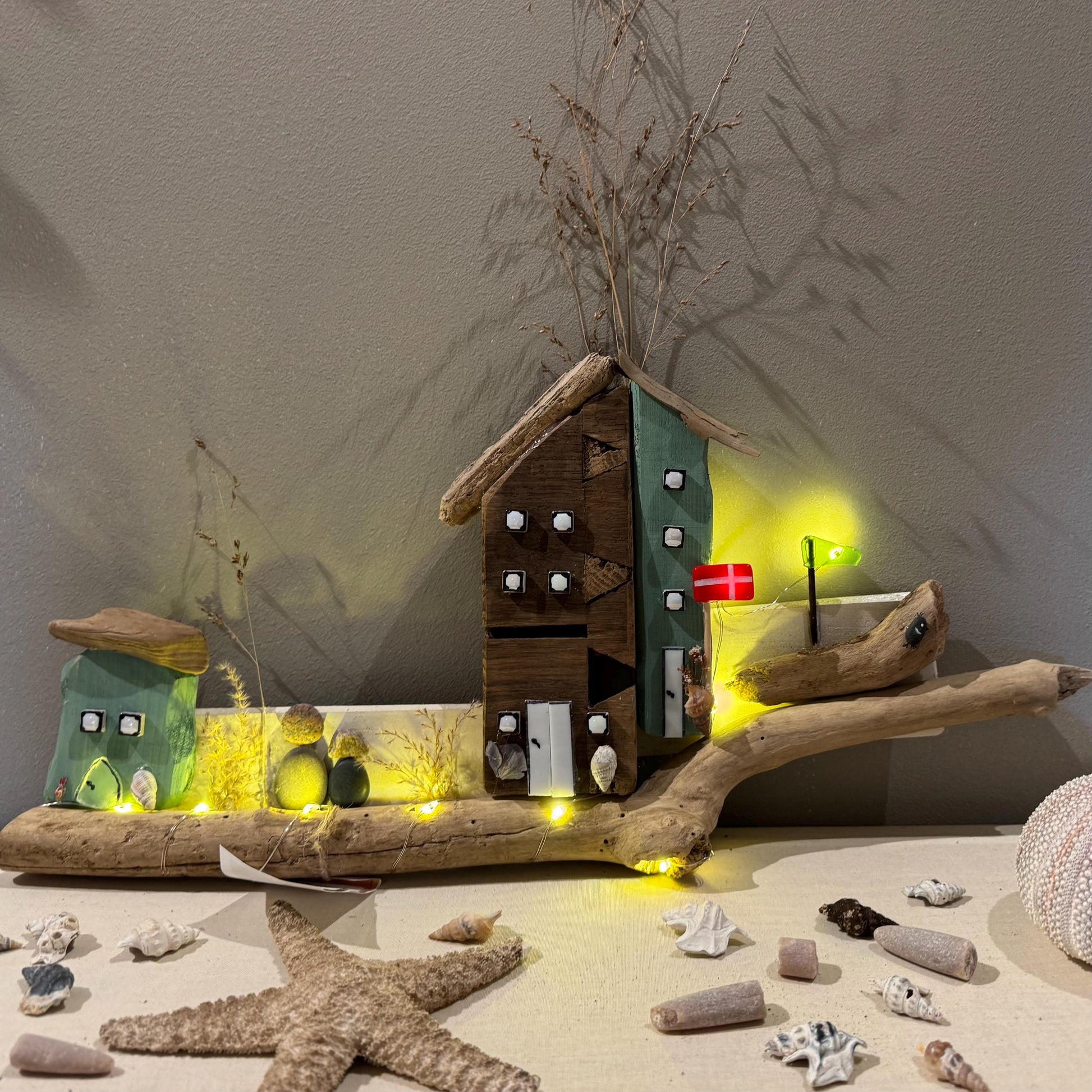 Treibholz Wanddeko Haus aus Treibholz mit kleinen Minihäusern und warmen LED-Lichtern auf heller Wand, maritime Dekoration.