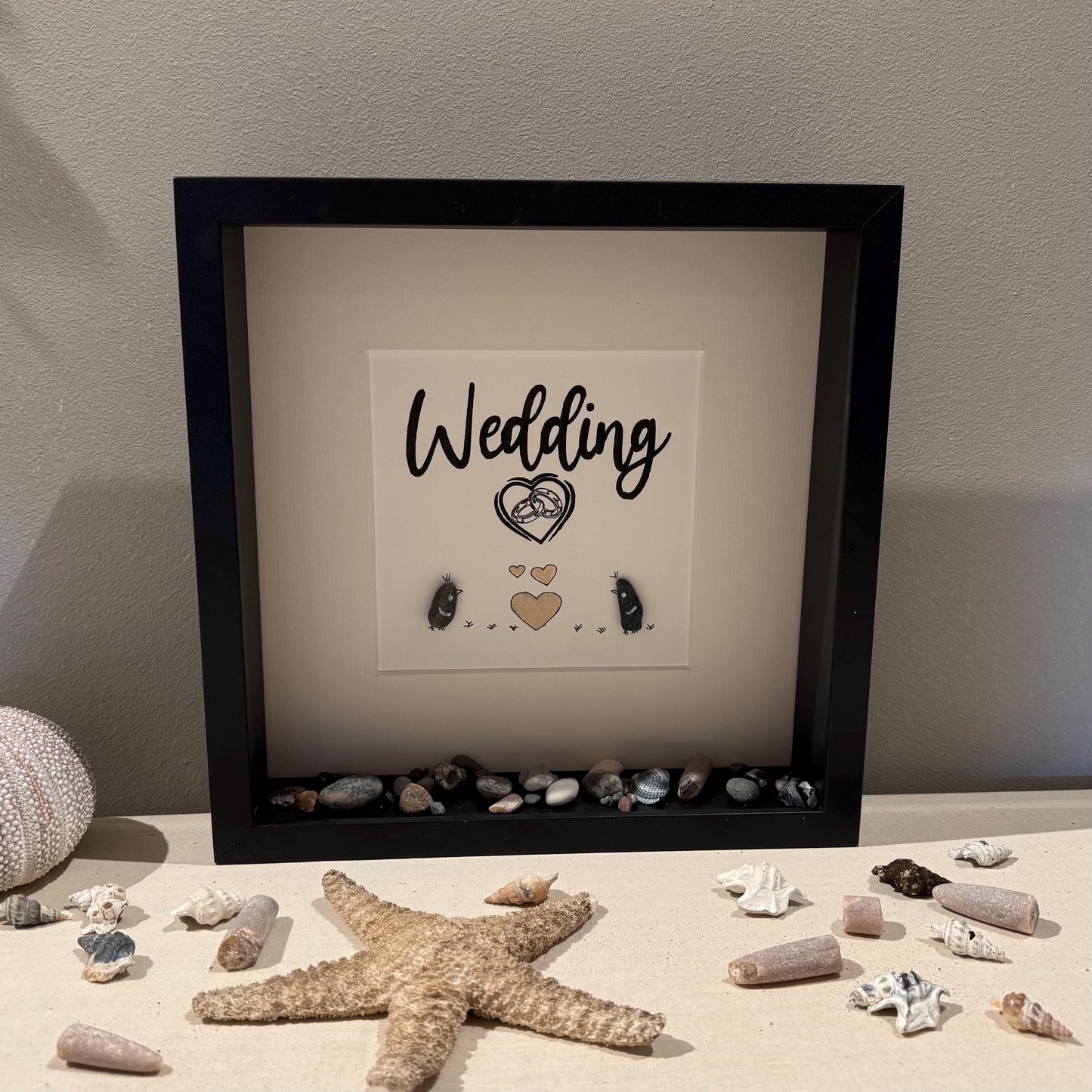 Ein gerahmtes Kunstwerk zum Thema Hochzeit mit dem Wort "Wedding" in Schriftform. Die Kunst zeigt zwei Steinmännchen
