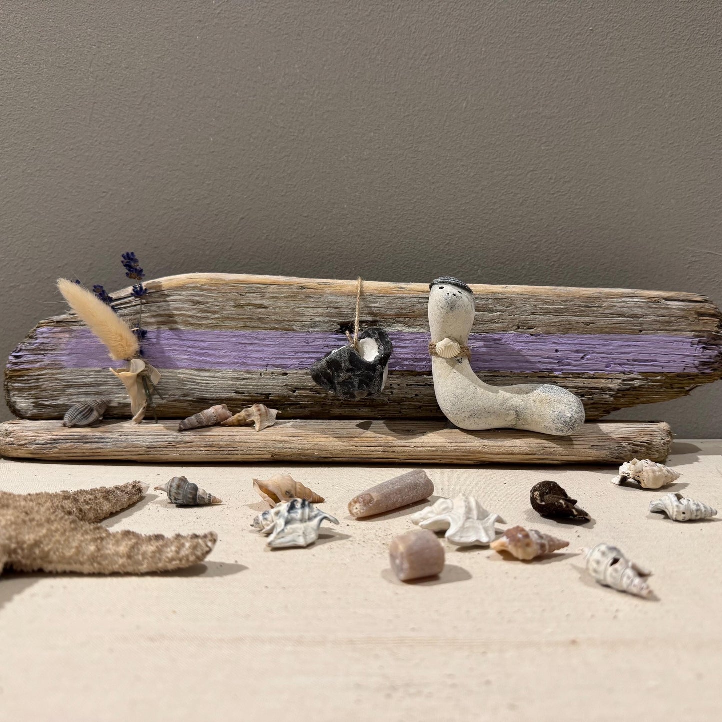 Maritime Treibholz Deko mit kleiner Steinfigur und Muscheln, liebevoll arrangiert auf Treibholz vor einer gemütlichen Couch.